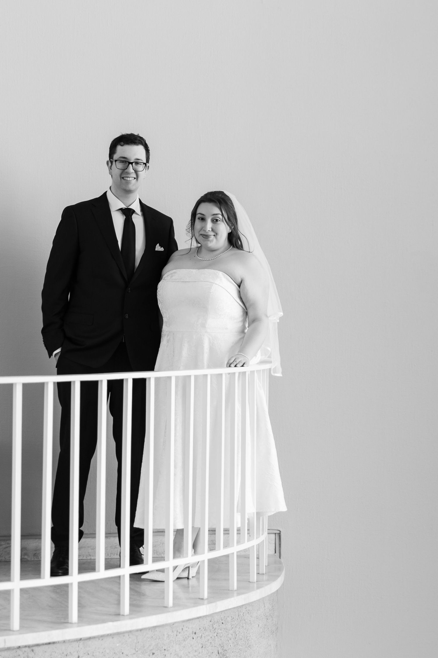 Black and white photo of bride and groom standing together on curved balcony railing smiling.