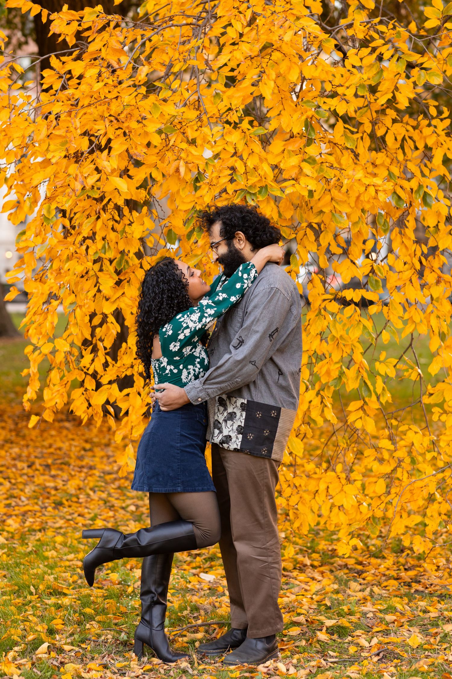 A couple embraces and gazes at each other under a golden autumn tree with fallen leaves on the ground.
