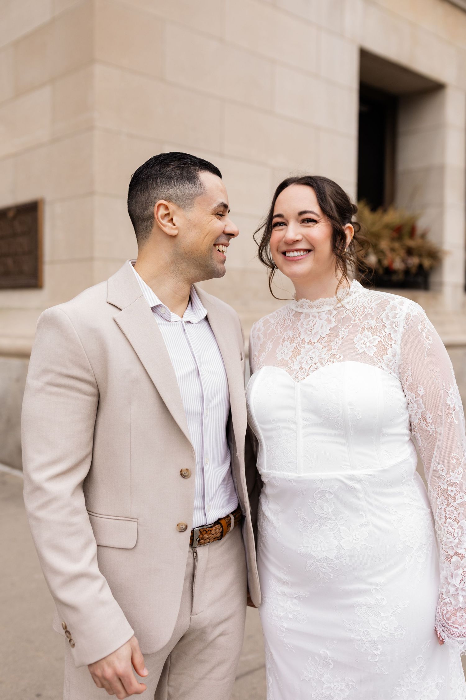 Smiling couple in wedding attire sharing a tender moment outside a stone building.