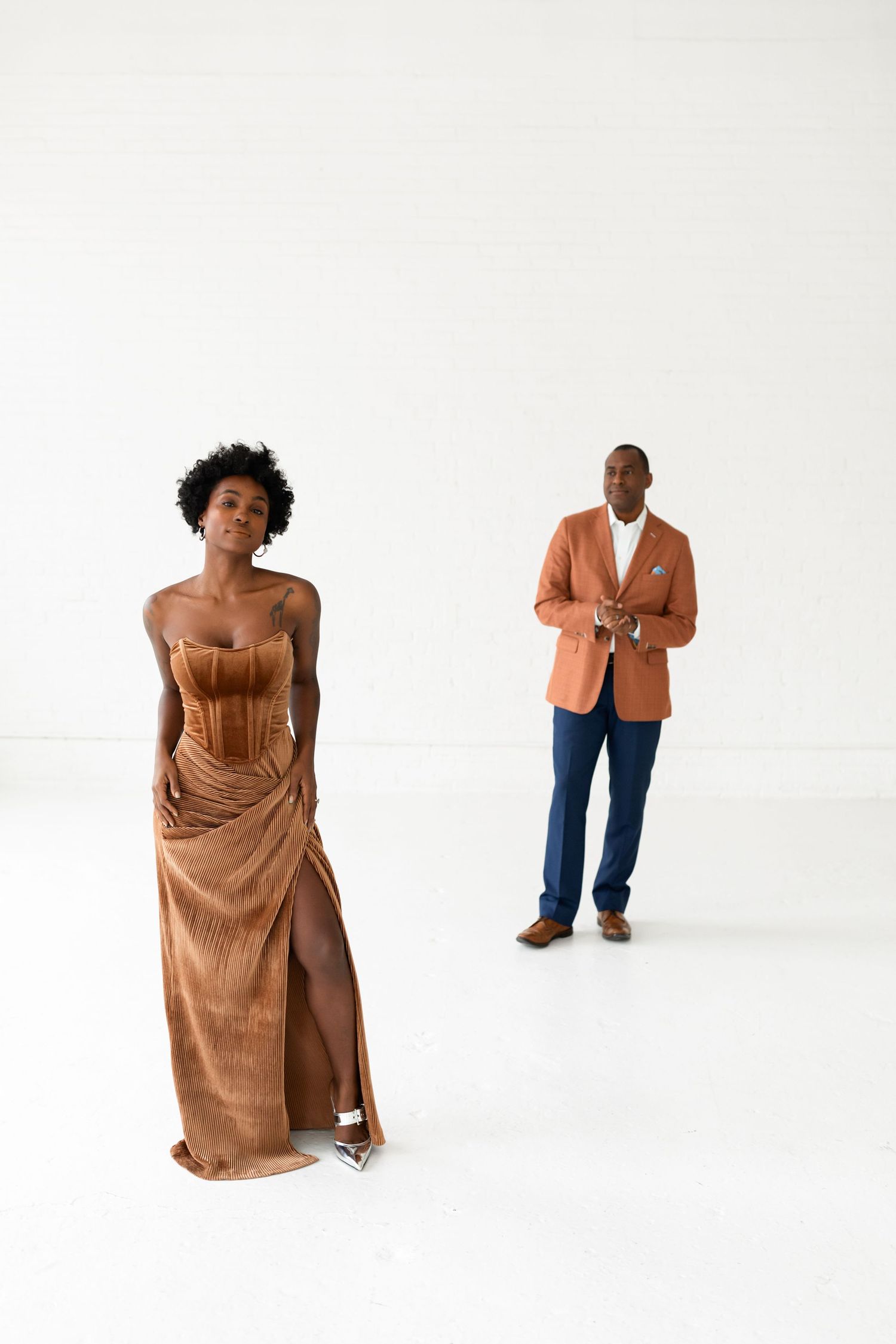 A woman in a gold velvet gown poses in the foreground while her partner stands in an orange blazer behind.