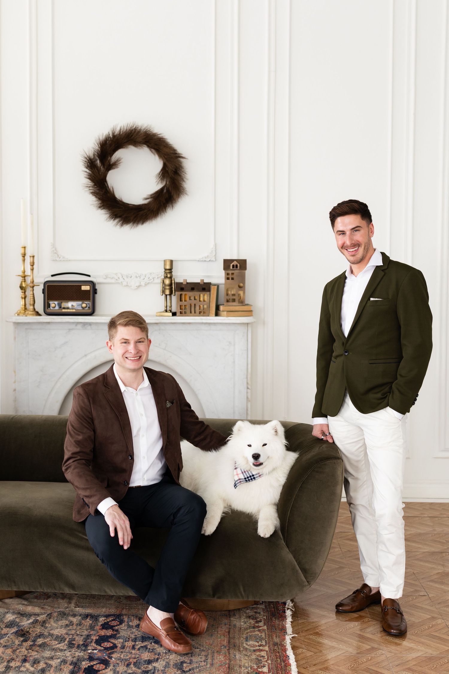 Two grooms in blazers pose with white Samoyed dog on green velvet sofa near elegant white fireplace mantel.