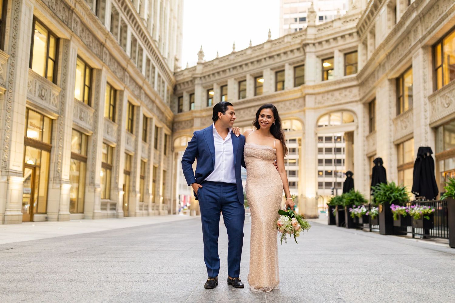 Bride in champagne sequin gown and groom in navy suit walking through ornate Chicago building arcade.