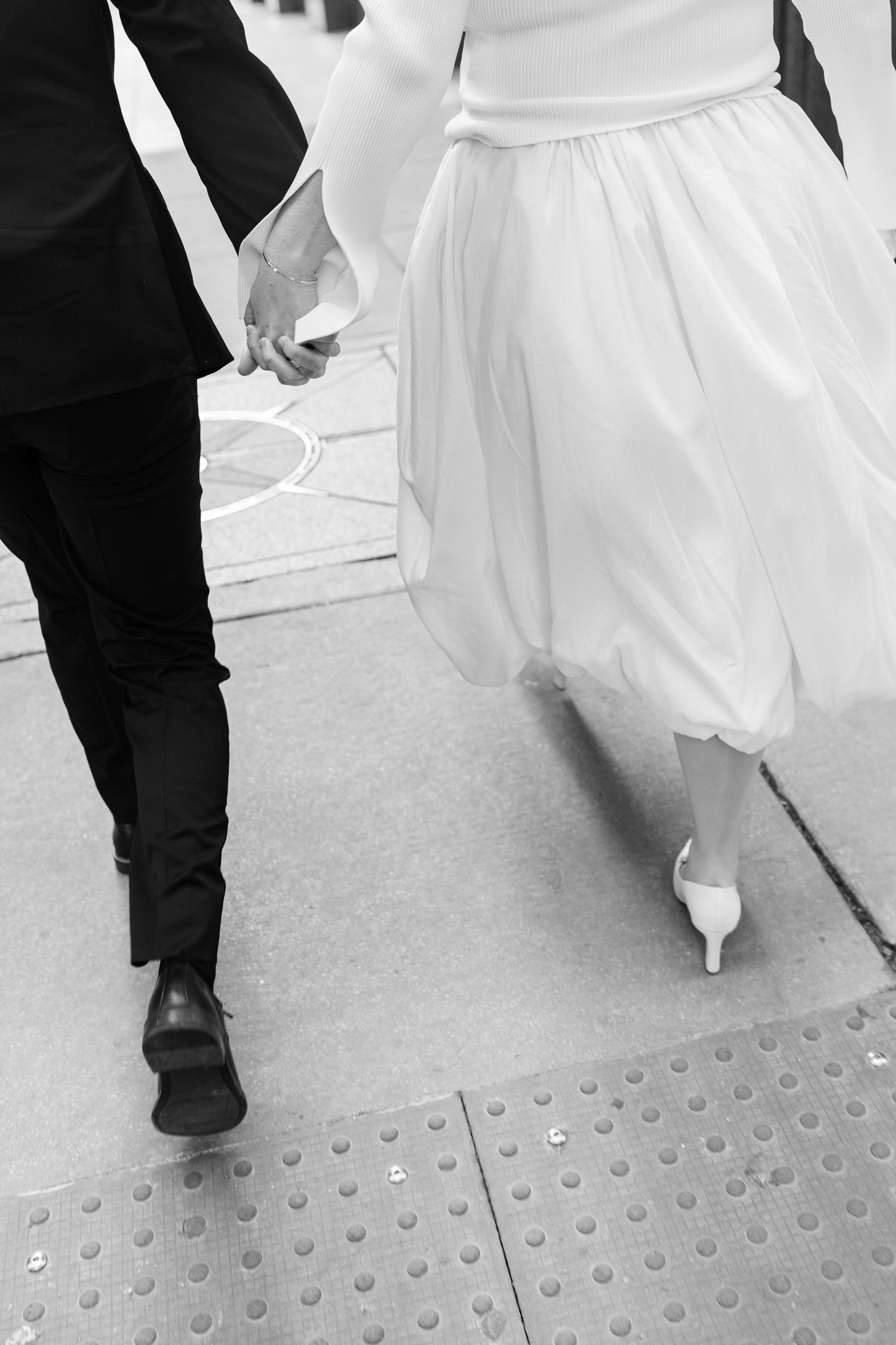 Black and white close-up of bride in white dress and groom holding hands while walking on sidewalk.