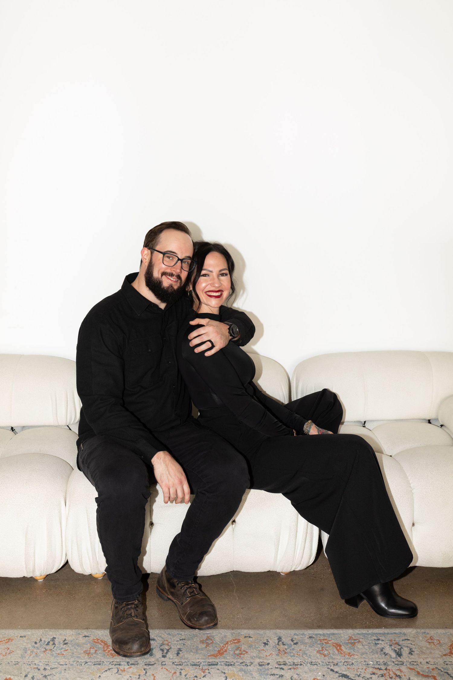 Happy couple in matching black outfits sitting close together on white sofa against clean white wall.