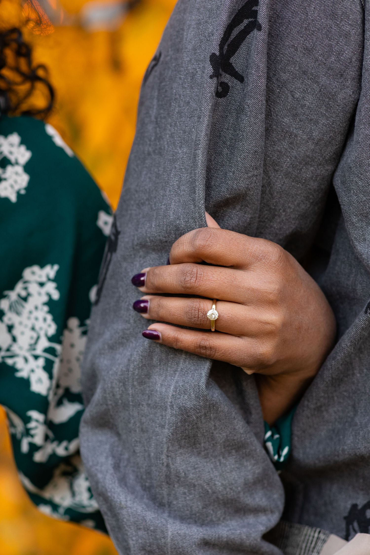 Close-up of a woman's hand with purple nails and a gold engagement ring holding her partner's arm.