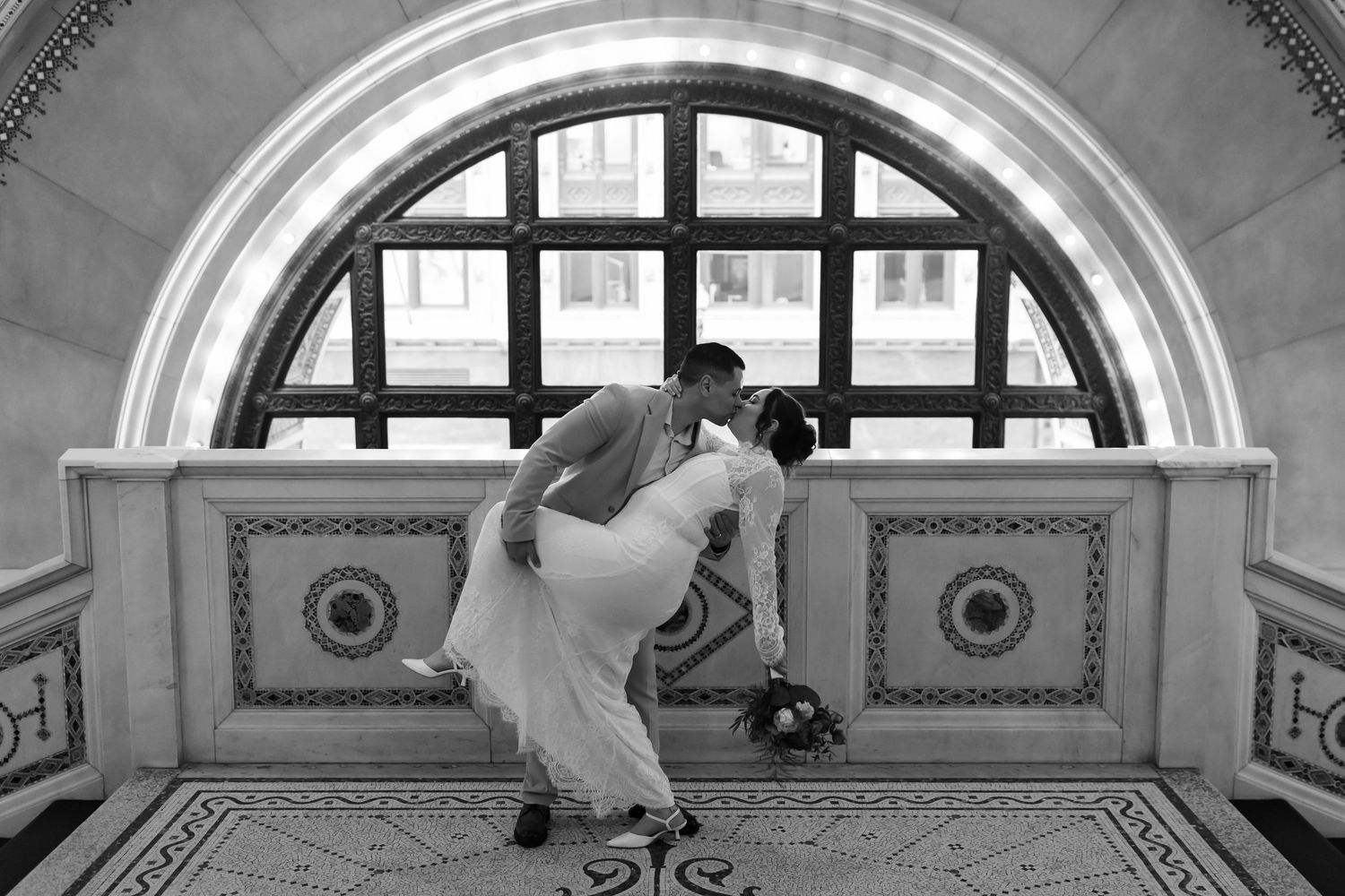 Black and white photo of a groom dipping his bride for a kiss under an ornate arched window.