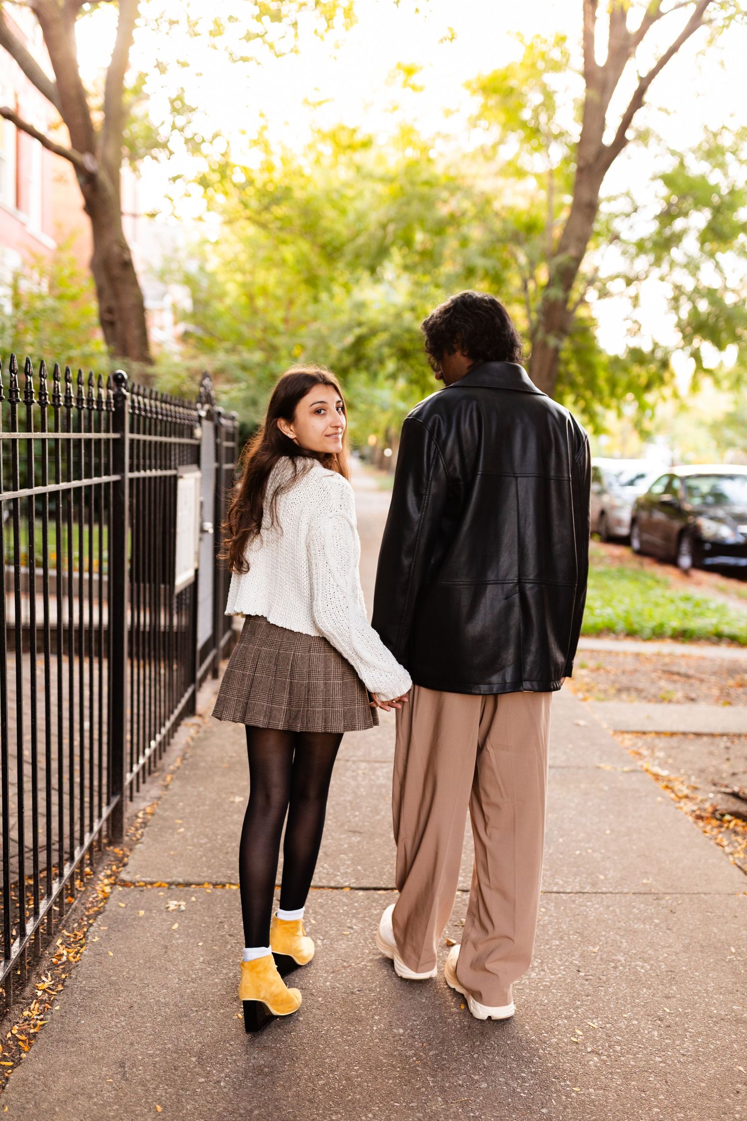 Young woman in yellow boots and plaid skirt looks back while holding hands with man in black leather jacket on sidewalk.