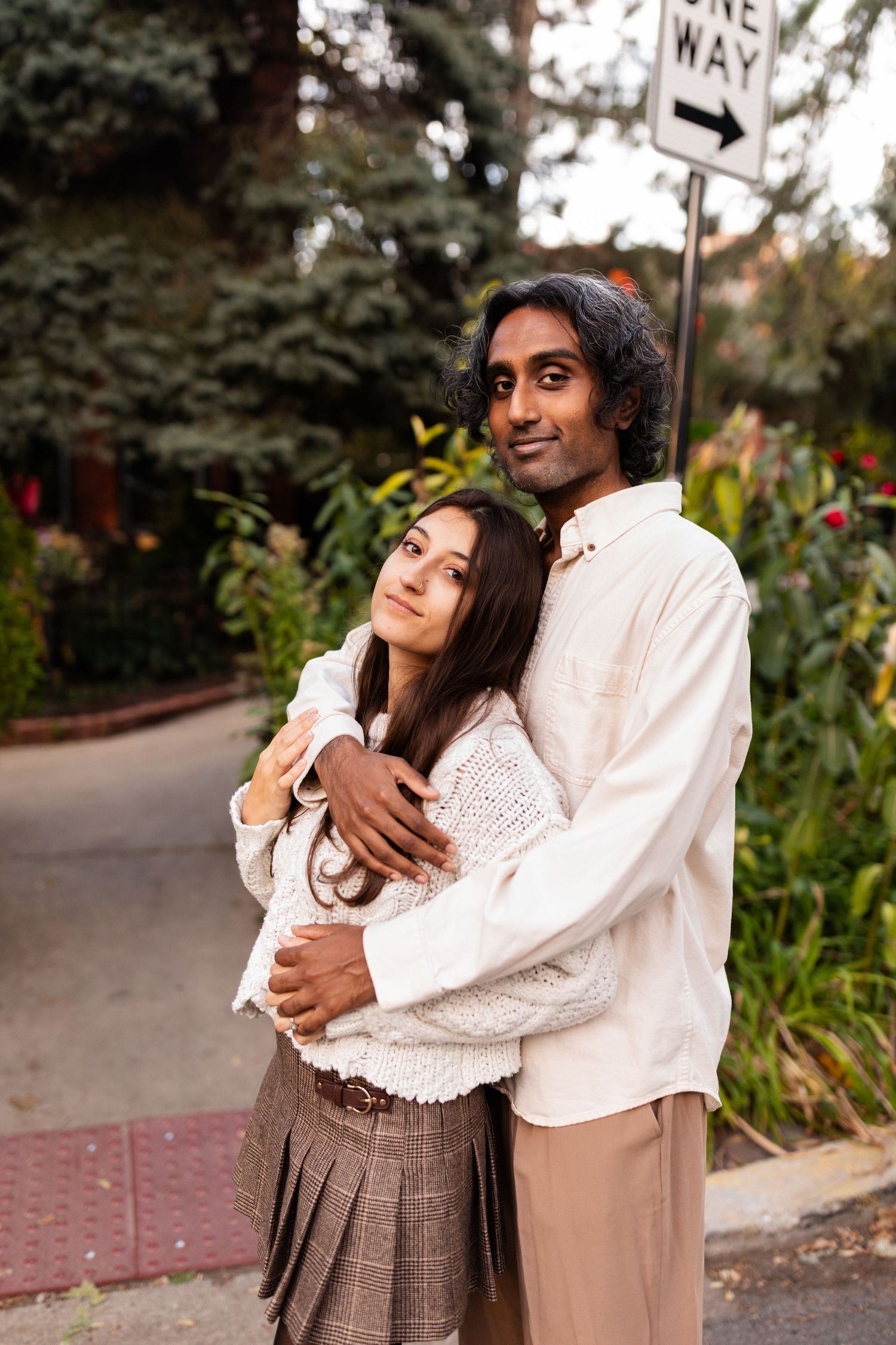 Couple hugging outdoors near a one-way street sign, surrounded by lush green foliage in autumn.