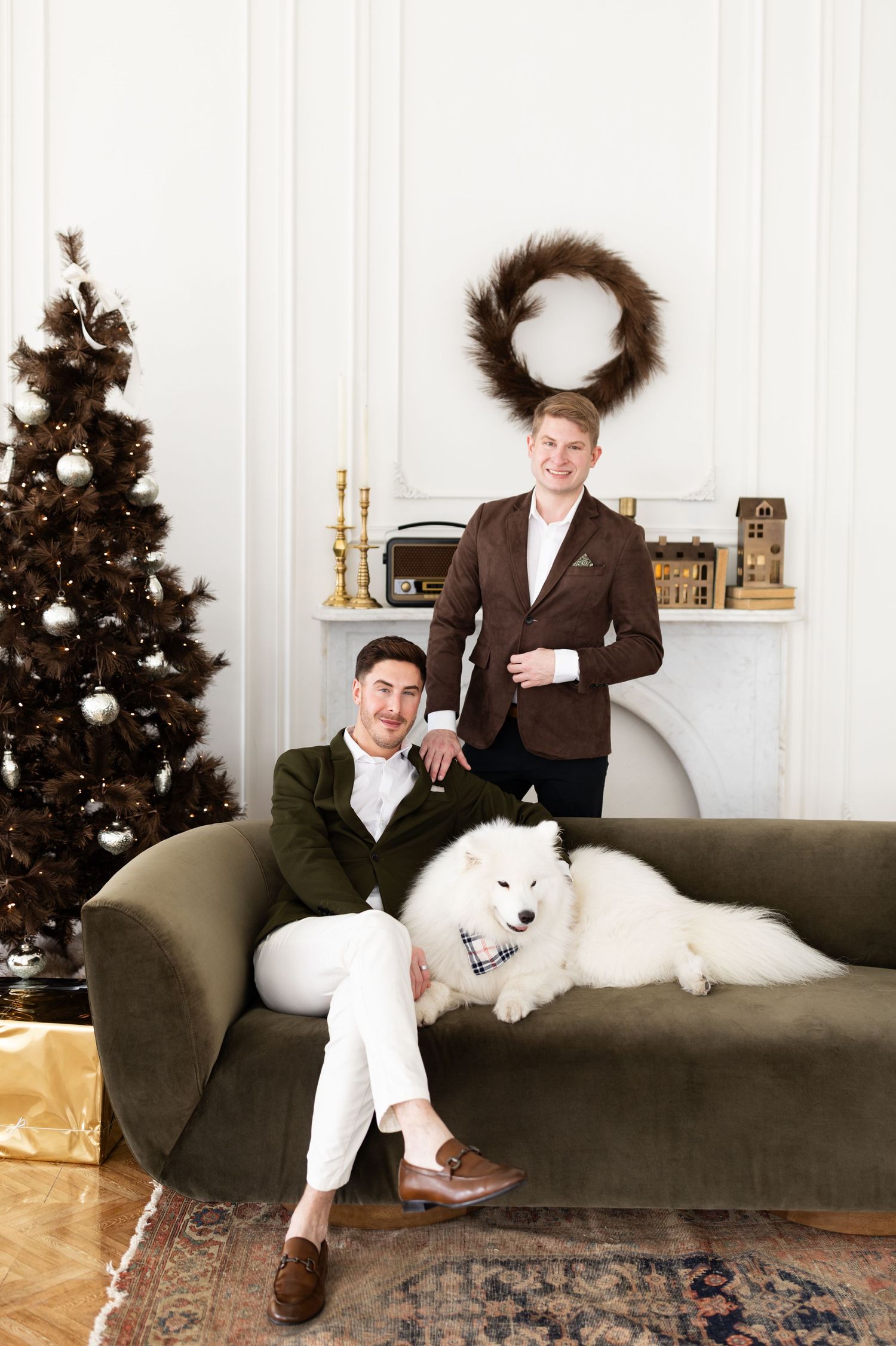 Two grooms relaxing on green velvet sofa with white Samoyed dog beside decorated Christmas tree and fireplace.
