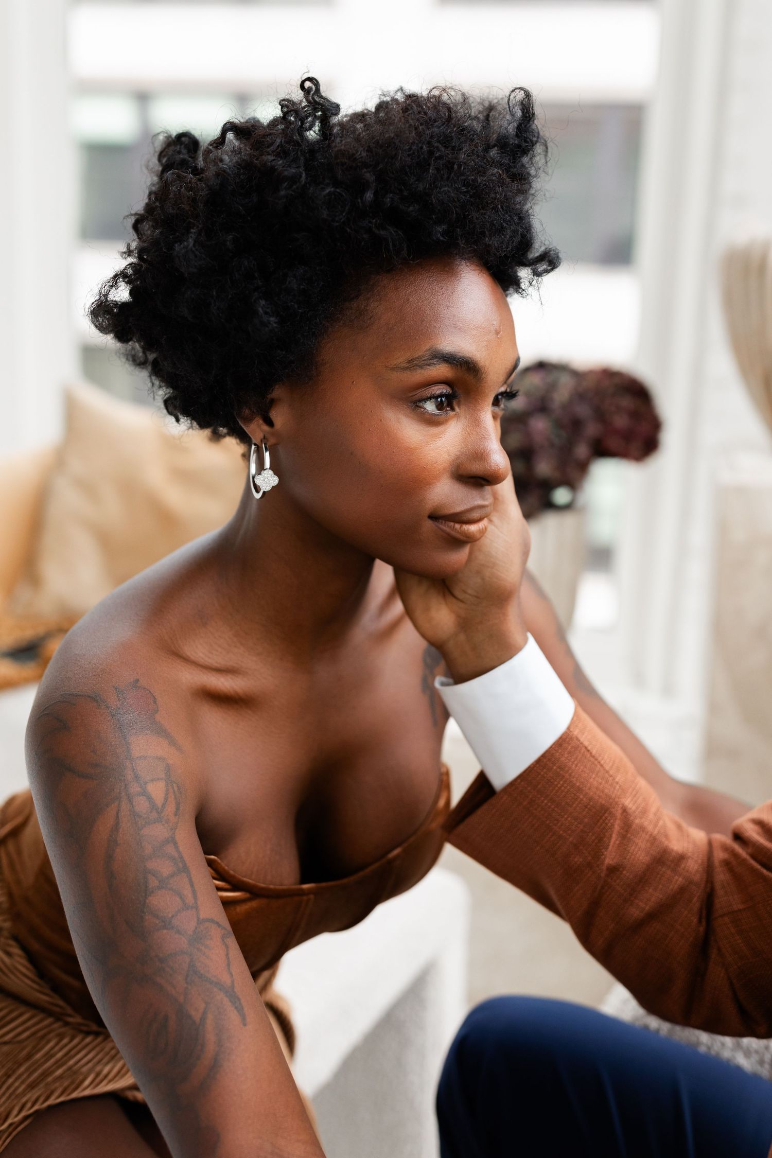 Beautiful Black woman with natural curly hair and tattoos in brown dress gazes thoughtfully while resting chin on hand.