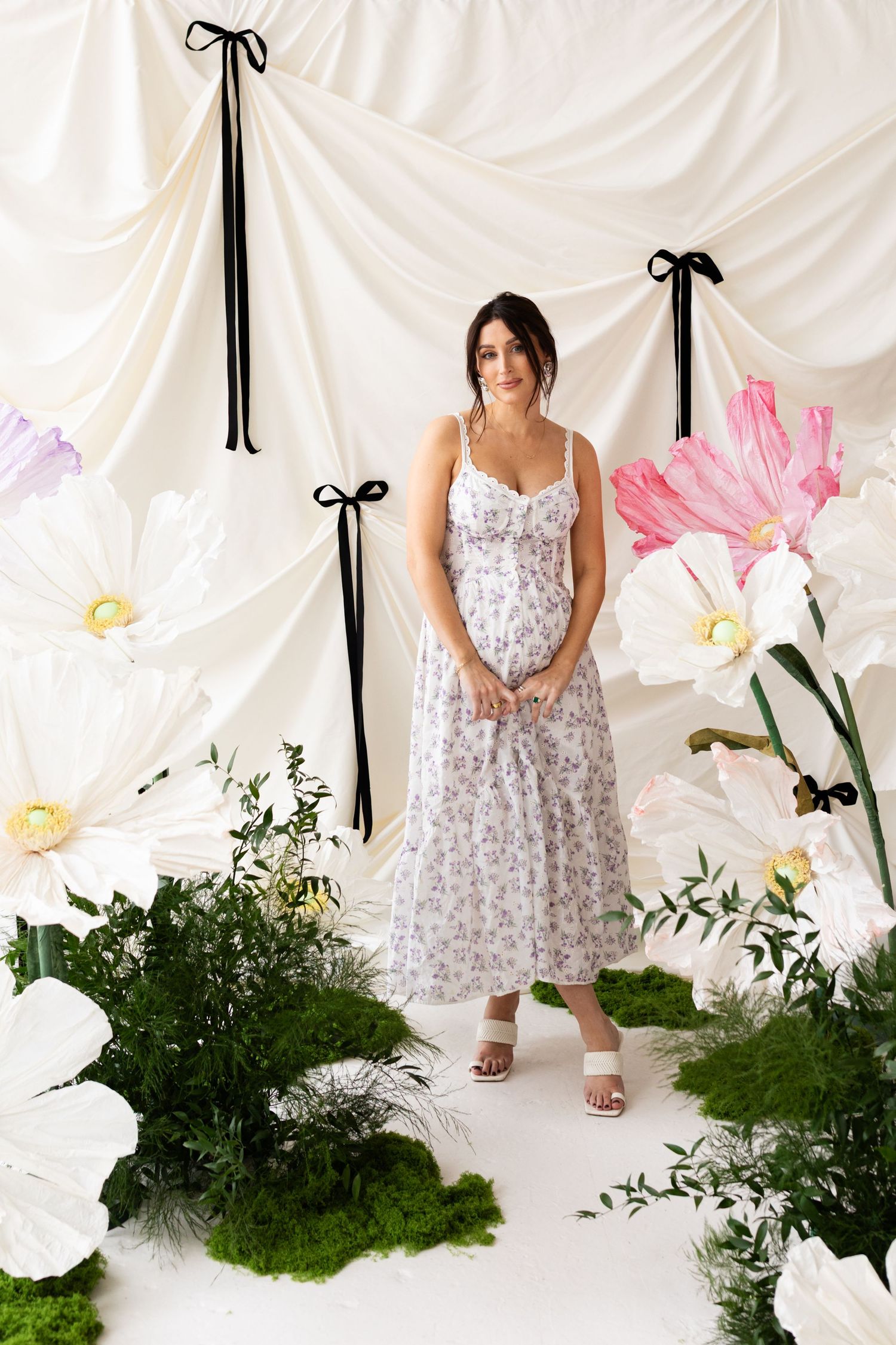 Pregnant woman in floral sundress stands among oversized paper flowers and lush greenery backdrop.