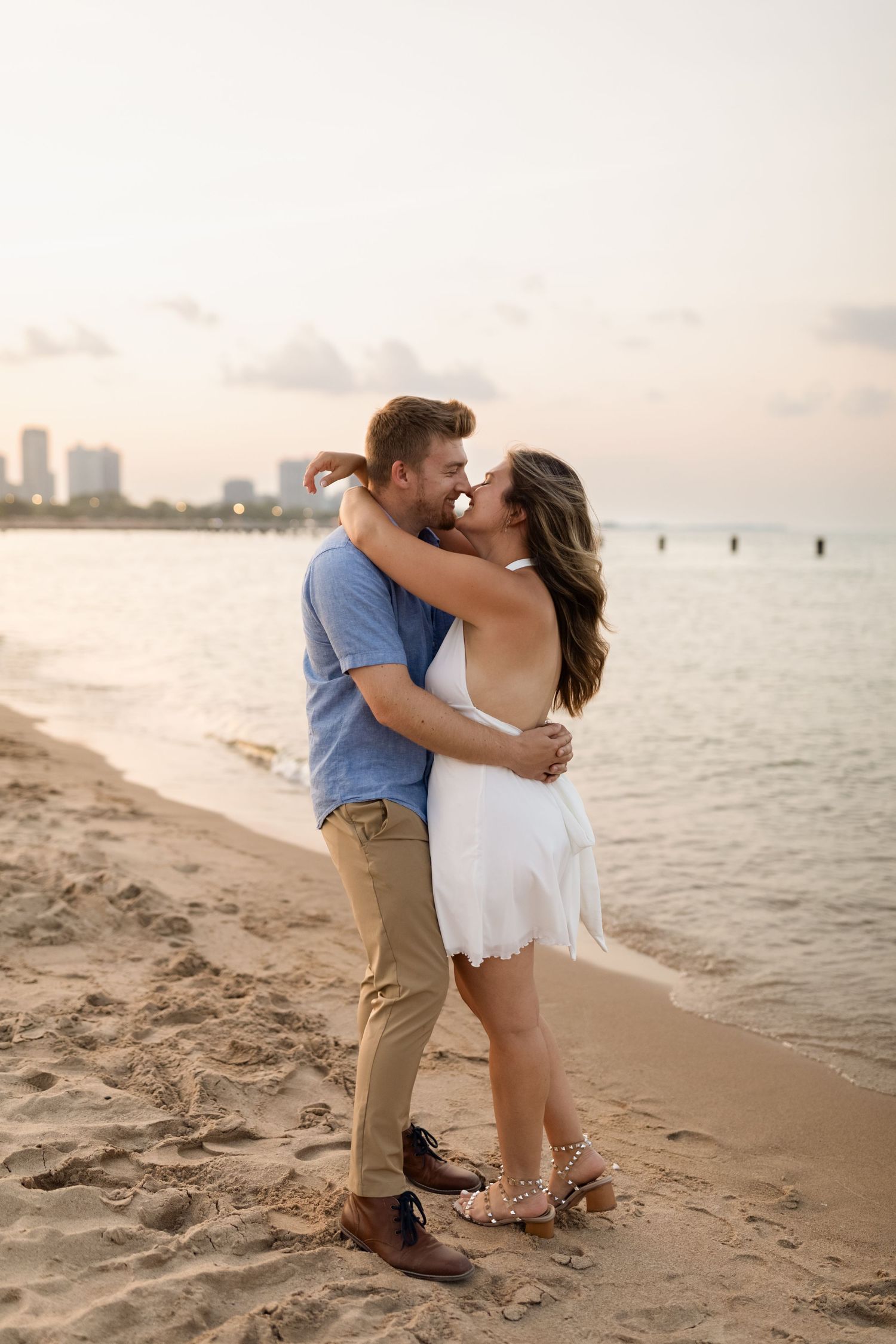 Couple embracing and almost kissing on a sandy beach at sunset with city skyline in background.