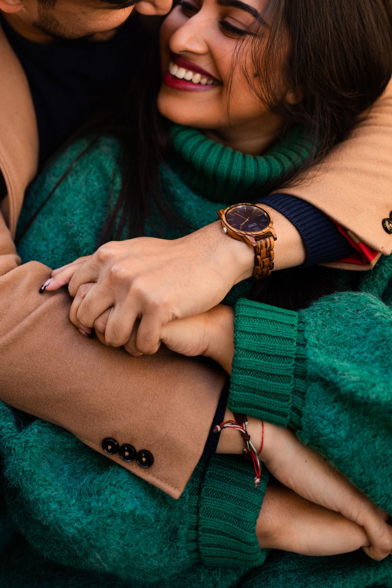 Close-up of couple hugging, woman in green turtleneck sweater laughing joyfully, man wearing wooden watch and bracelet.