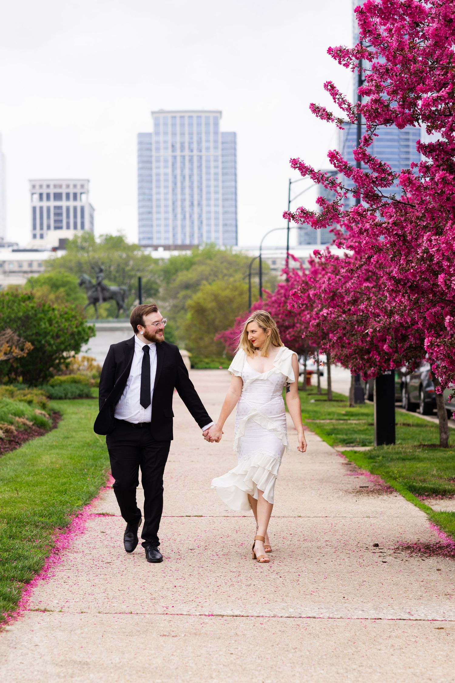 Newlyweds walk hand-in-hand along a path lined with vibrant pink flowering trees in Chicago.