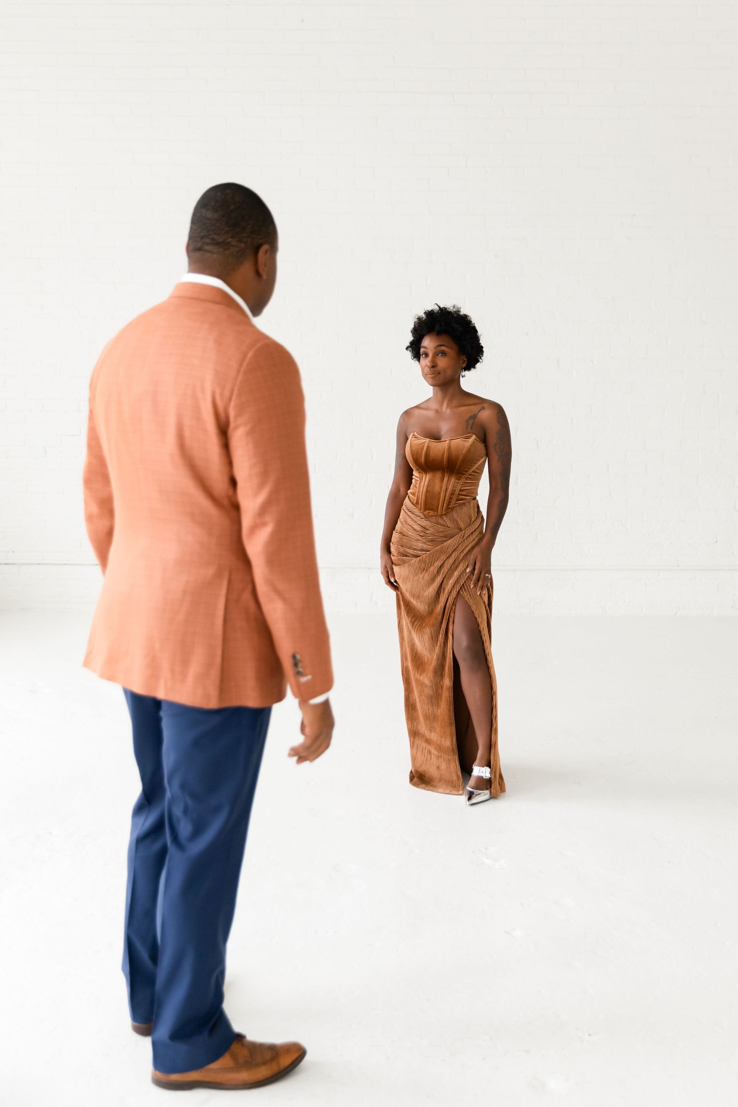 A stylish woman in a gold velvet gown walks toward a man in an orange blazer in a white studio.