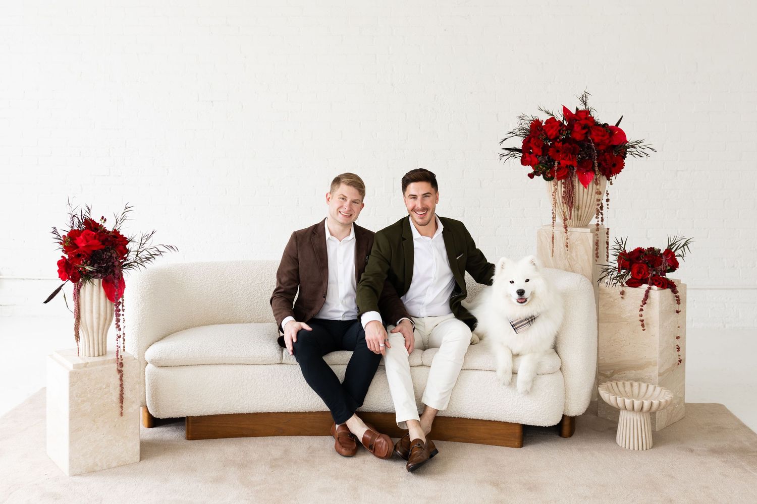 Two men and a white Samoyed dog posing on white sofa surrounded by red floral arrangements.