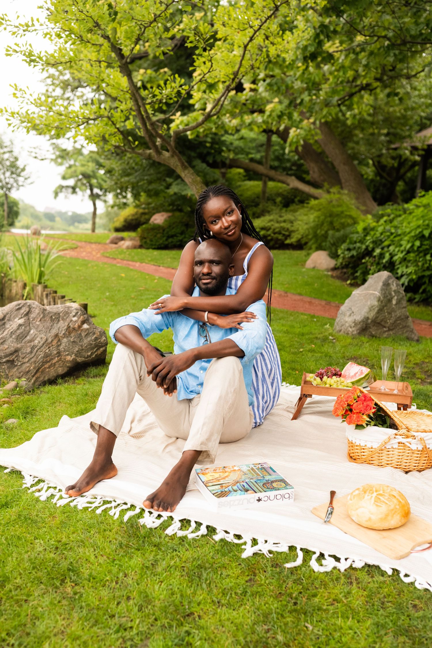 A couple enjoys a romantic picnic on a blanket in a lush green park, the woman embracing the man from behind.