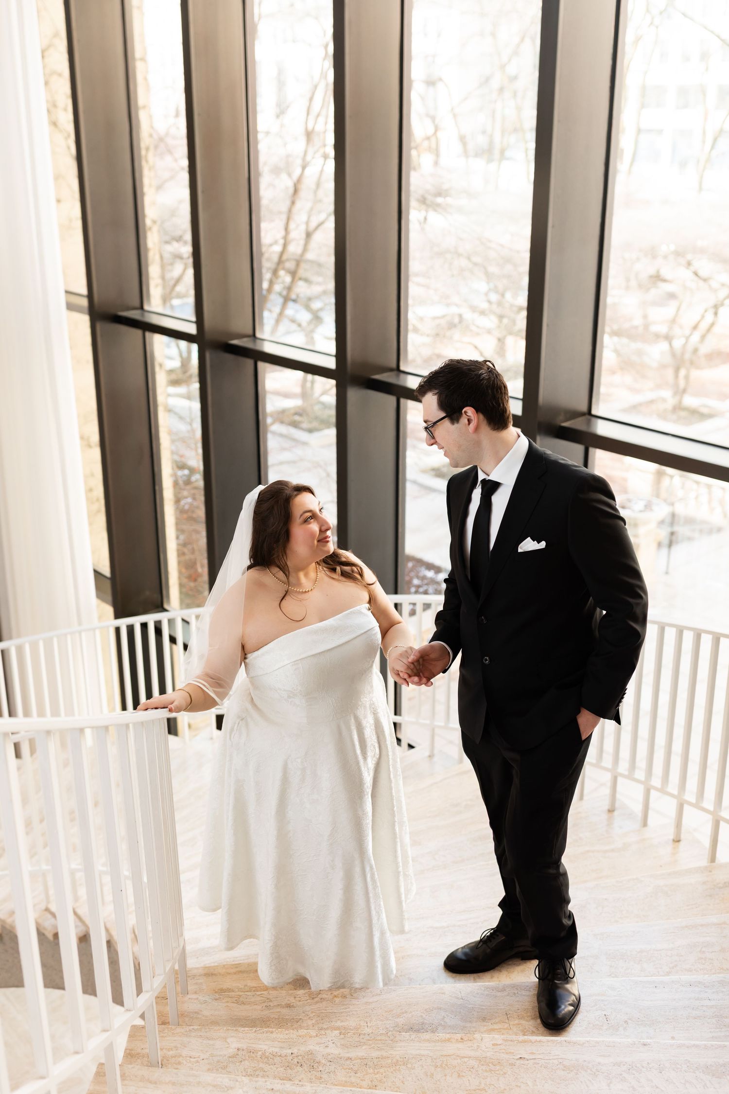 Bride and groom holding hands on elegant staircase with floor-to-ceiling windows in background.