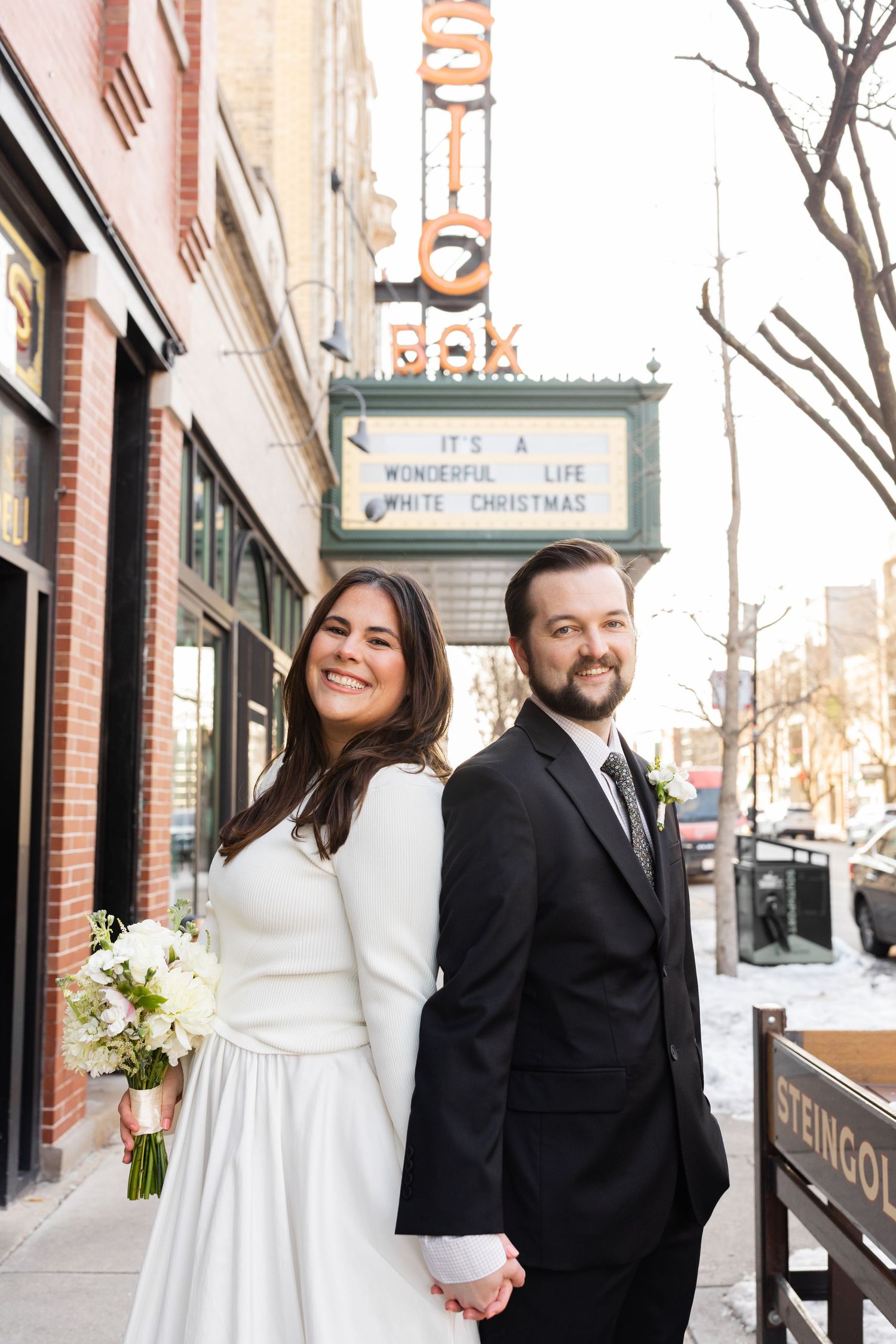 Bride and groom standing back to back holding hands outside Music Box Theatre with holiday marquee in Chicago.