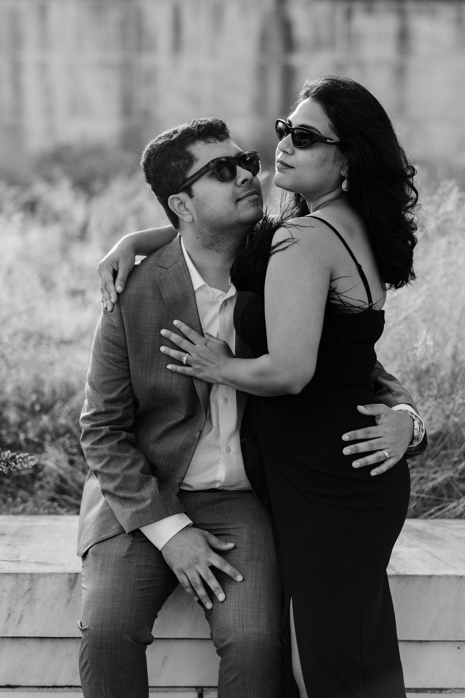 Black and white photo of an elegant couple in sunglasses sharing a romantic moment on a marble ledge.