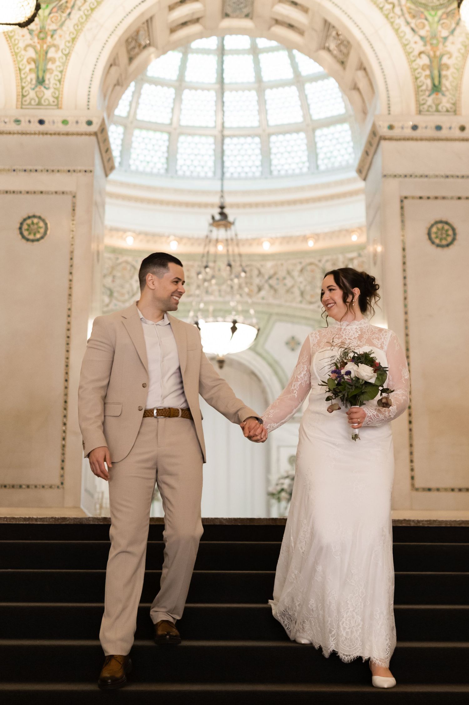 Bride in lace gown and groom in tan suit holding hands on grand staircase of elegant building.