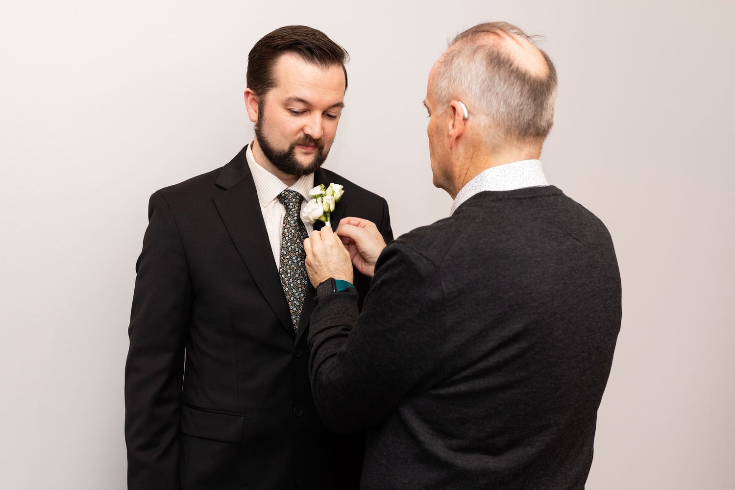 Older man pins white boutonniere on bearded groom in black suit before wedding ceremony.