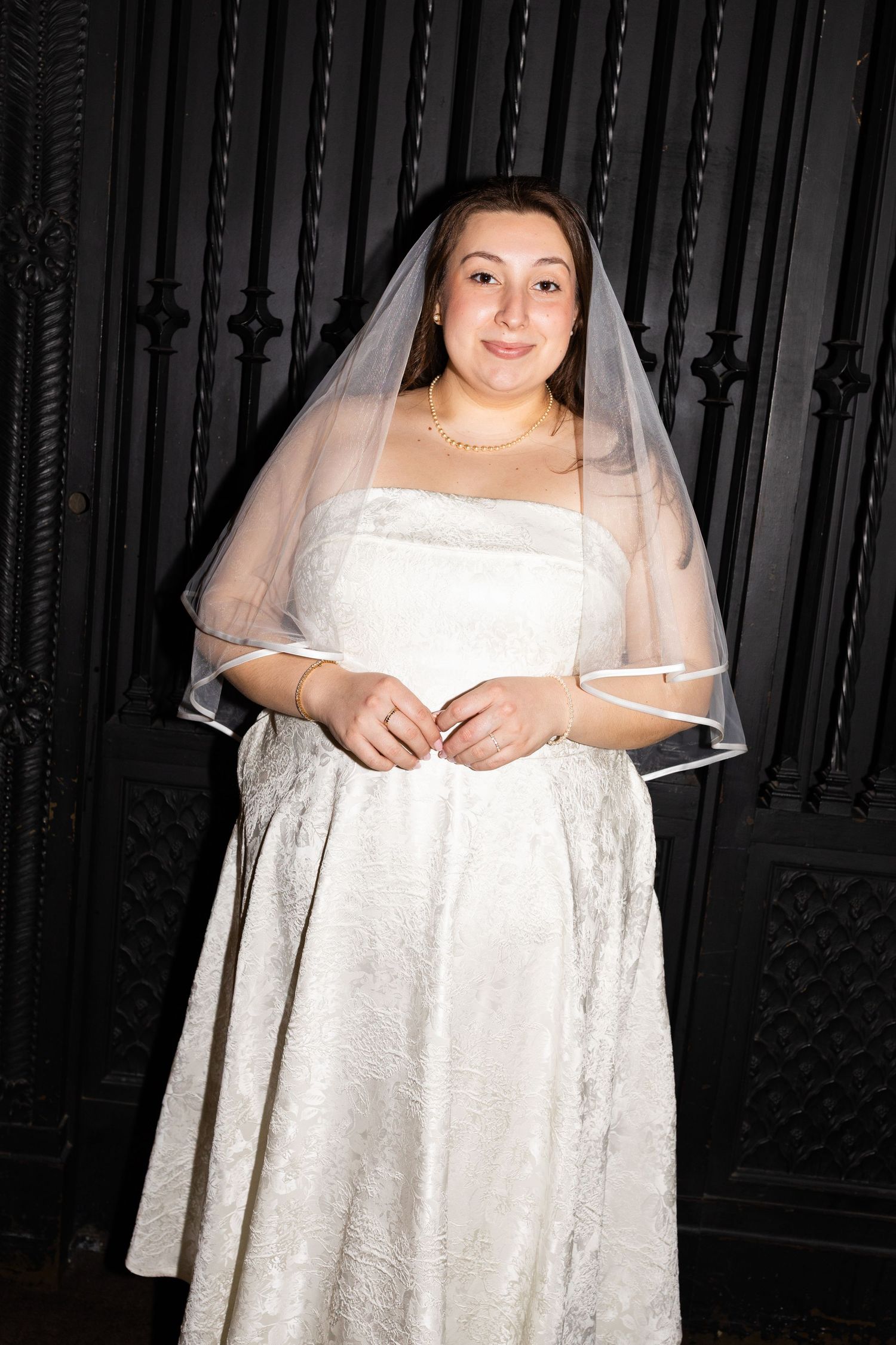 Bride in white lace gown and veil poses in front of decorative black iron gate.