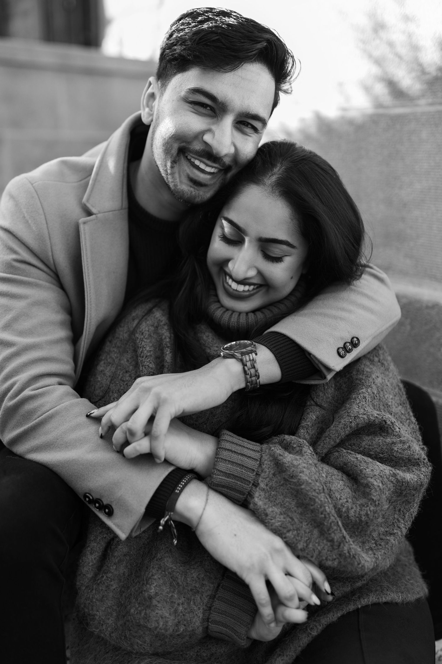Black and white photo of joyful couple hugging, man in blazer laughing, woman in cozy sweater smiling with eyes closed.