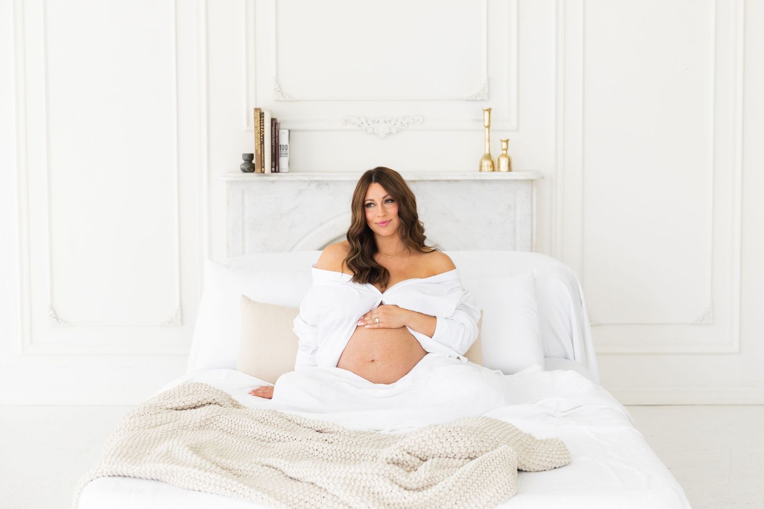 Pregnant woman sitting on white bed wrapped in white sheets in bright minimalist bedroom.