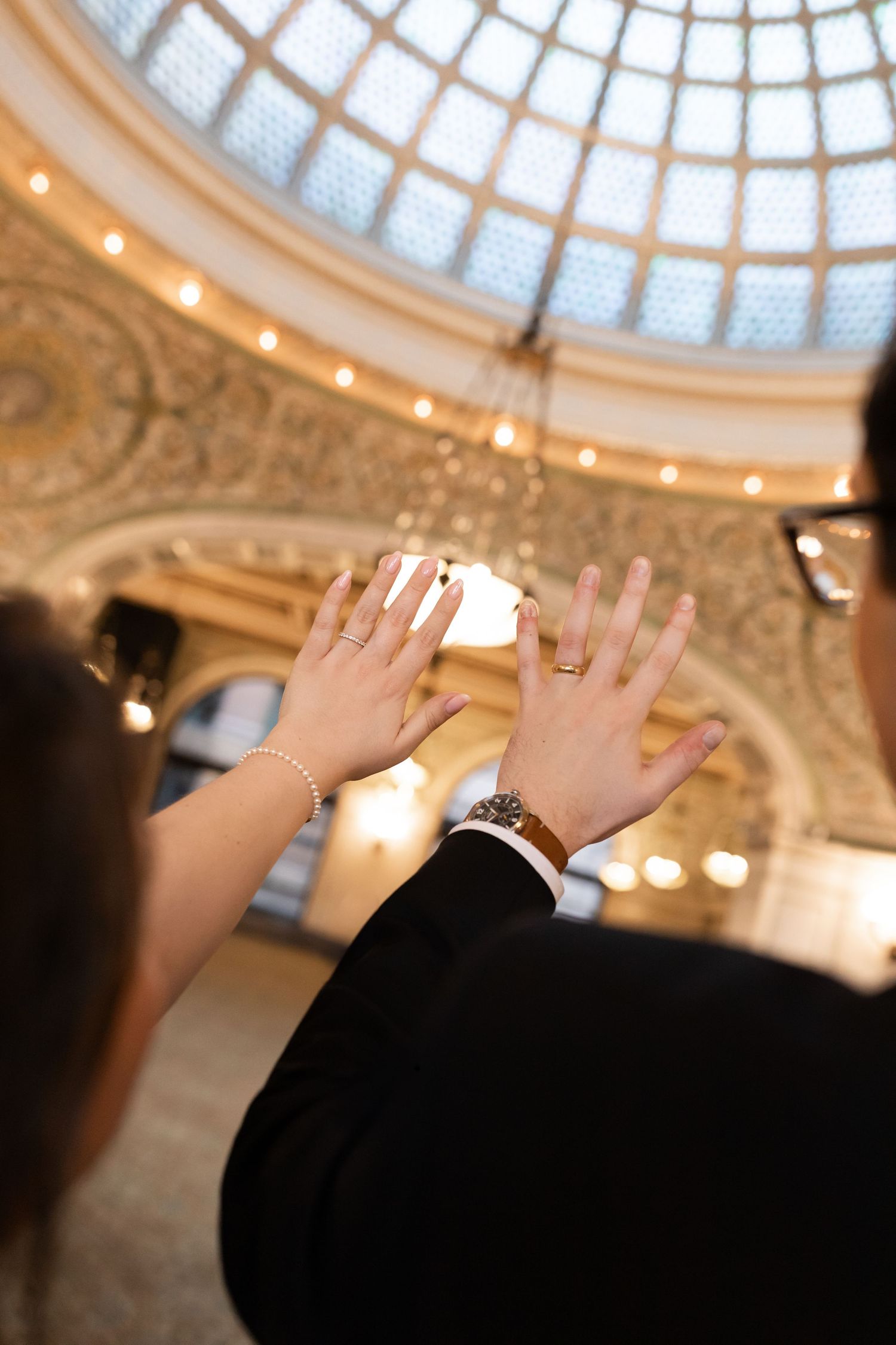 Couple raises hands showing wedding rings beneath stunning domed stained glass ceiling indoors.