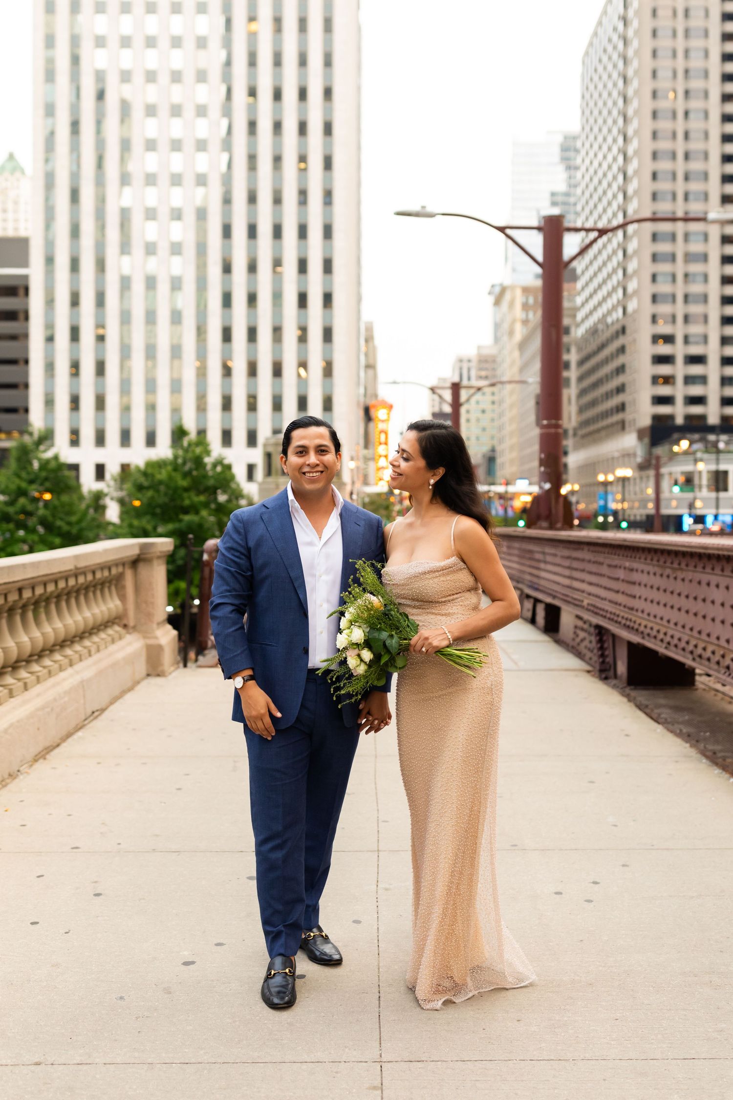 Elegant newlywed couple posing on Chicago bridge, bride in gold gown holding lush green bouquet.
