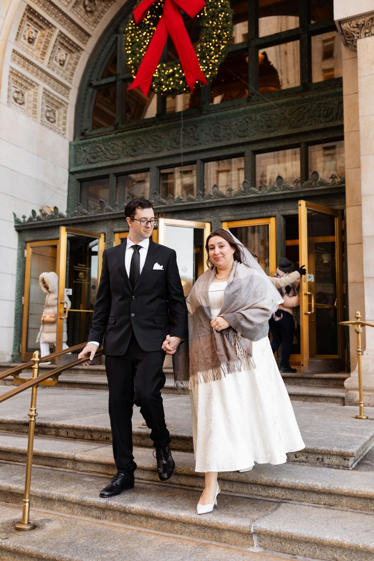 Bride and groom descend steps of elegant building entrance adorned with Christmas wreath.