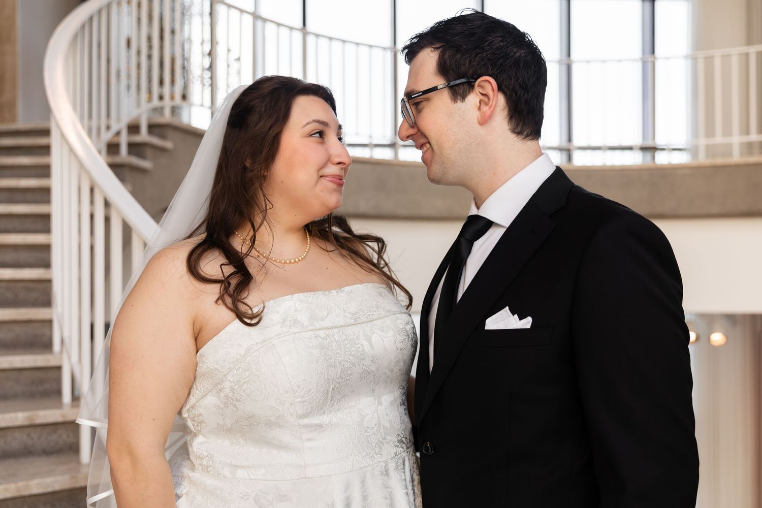 Bride and groom gazing at each other lovingly near a spiral staircase in a bright venue.