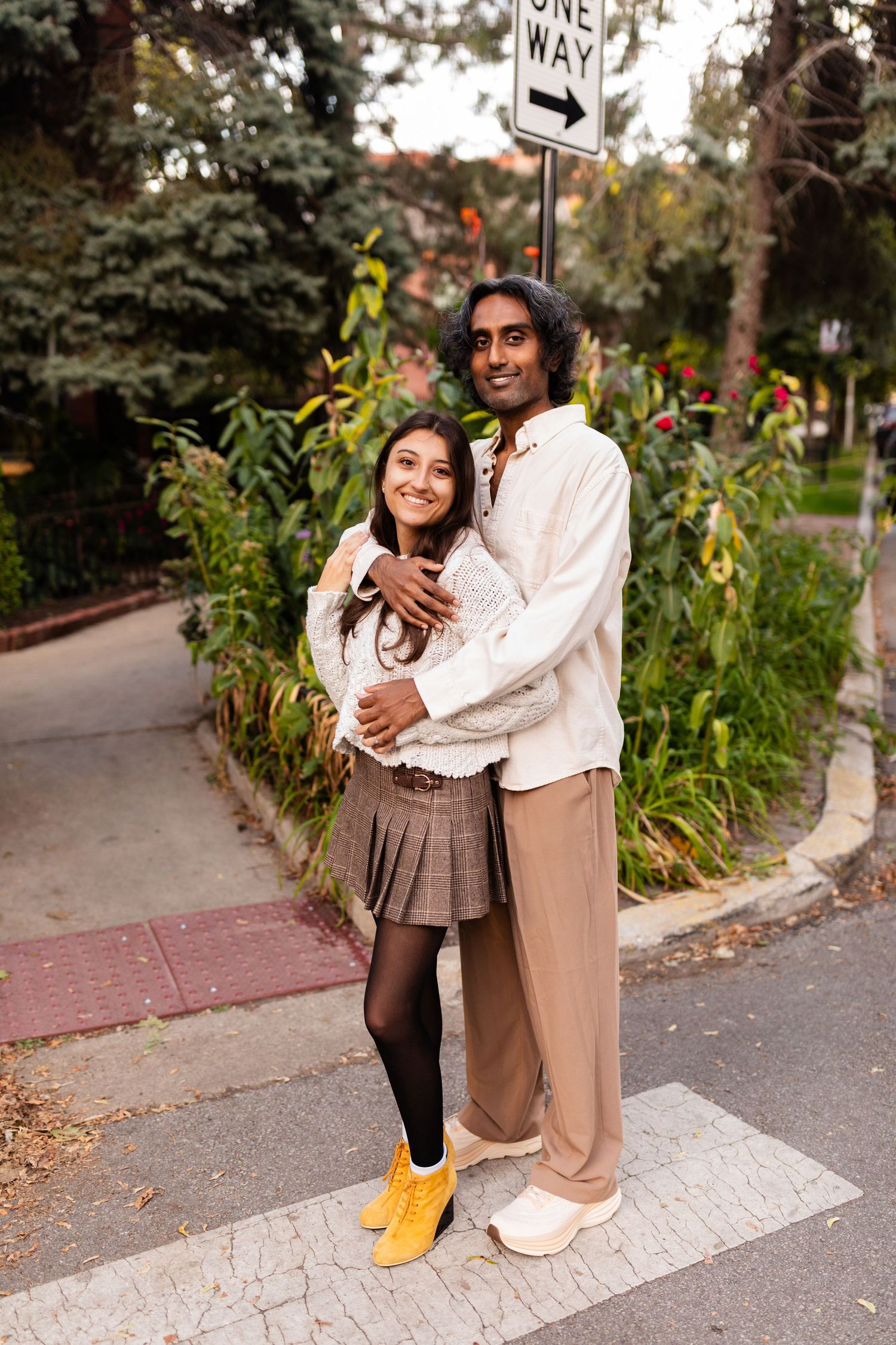 Smiling interracial couple embracing on urban crosswalk, woman in white sweater and plaid skirt with yellow boots.