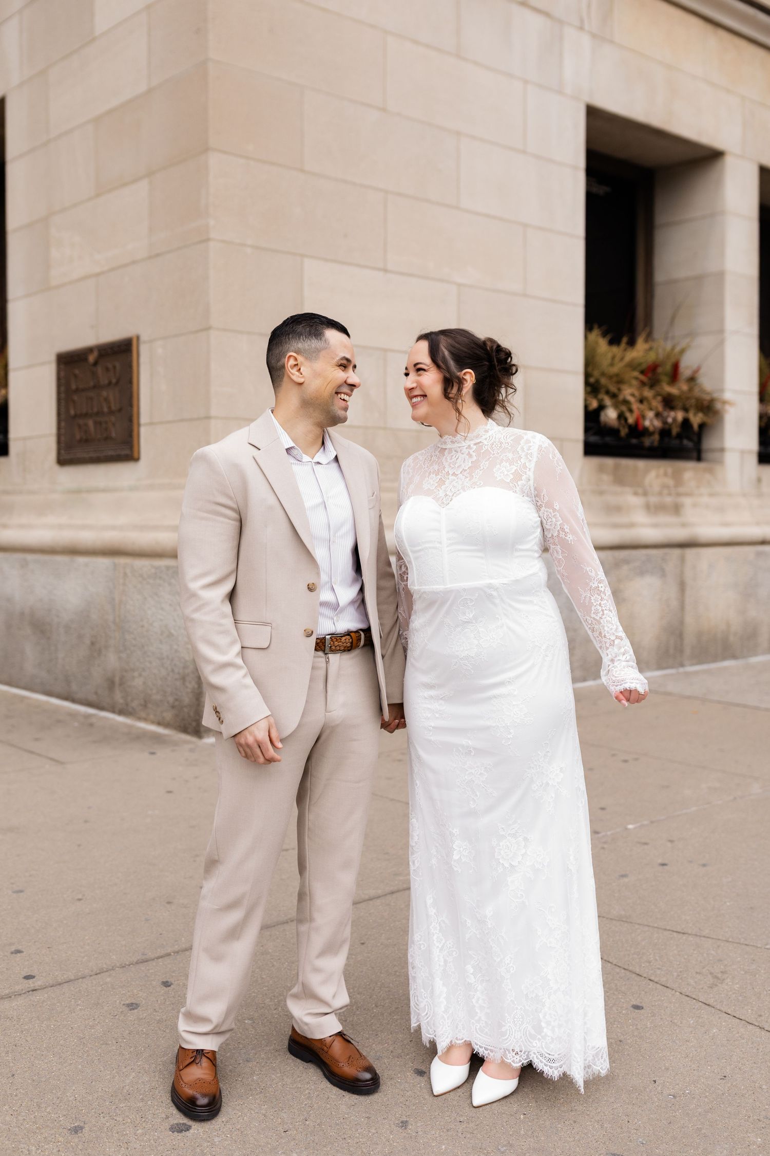 Bride in long-sleeve lace gown and groom in tan suit holding hands and smiling outside stone building.