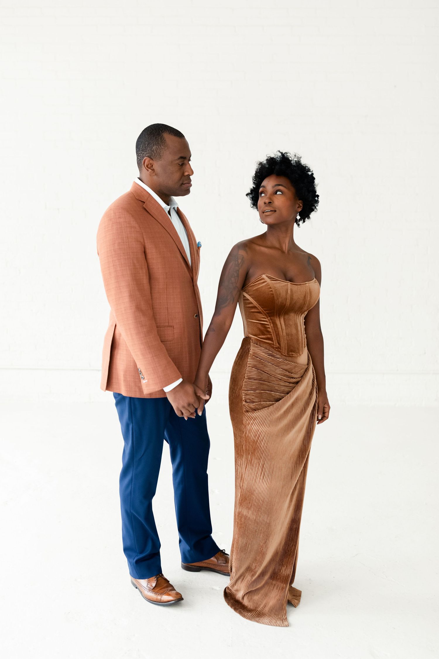 An elegant couple holding hands and gazing at each other, dressed in formal attire against a white backdrop.