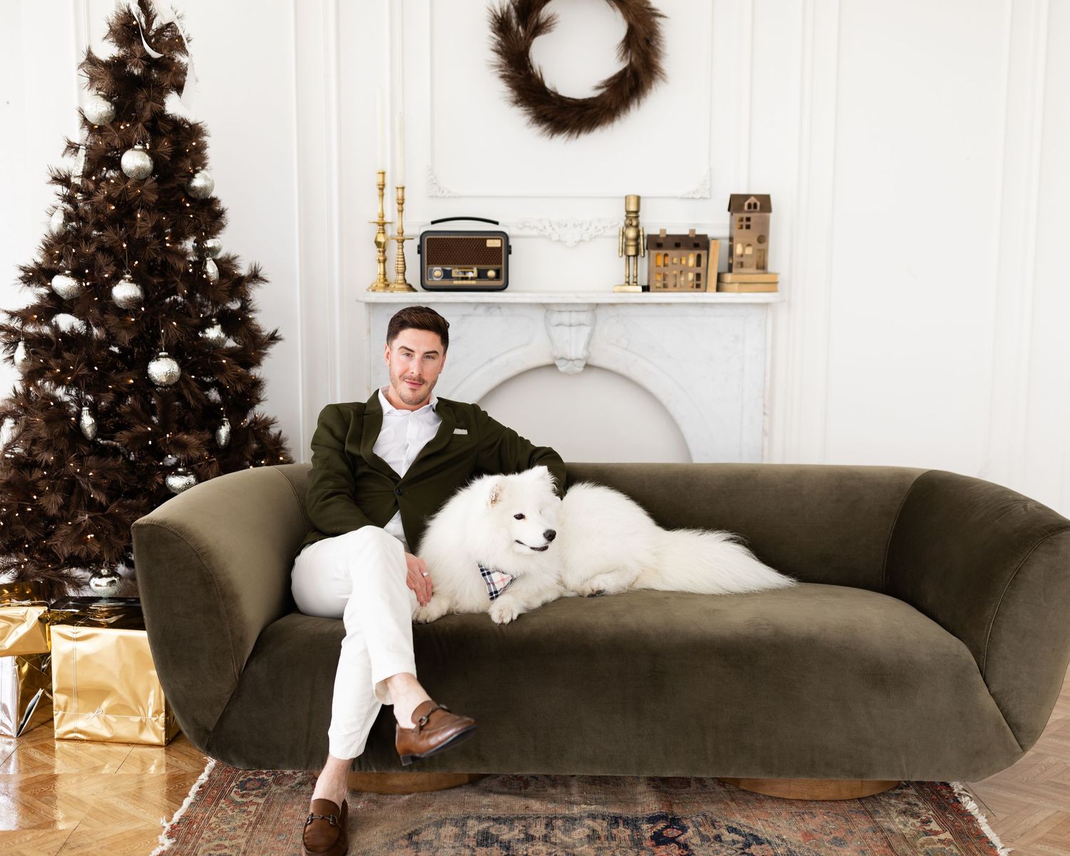 Man in green blazer relaxes on olive velvet sofa with white Samoyed dog near Christmas tree.