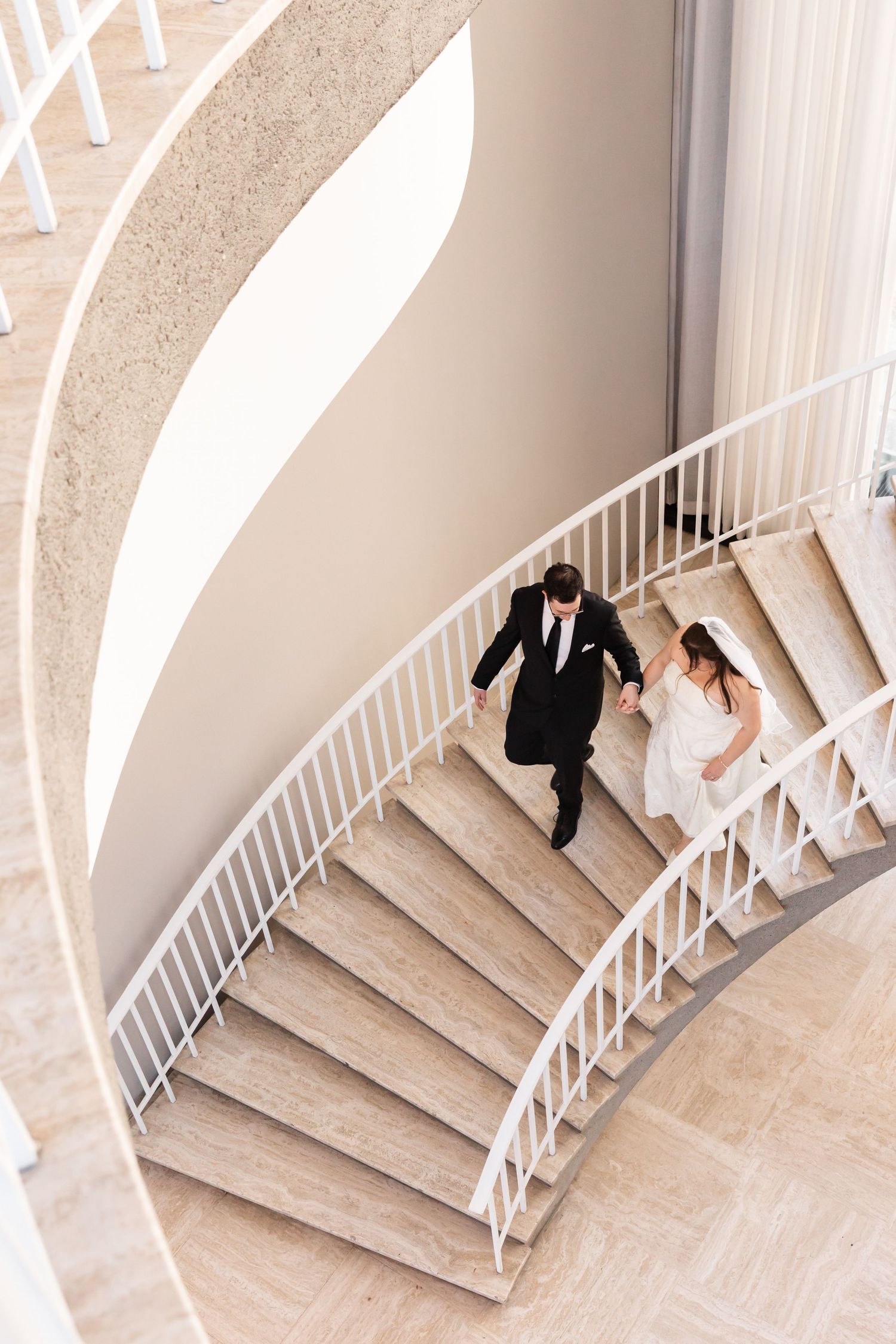 Aerial view of bride and groom holding hands while ascending an elegant spiral staircase together.