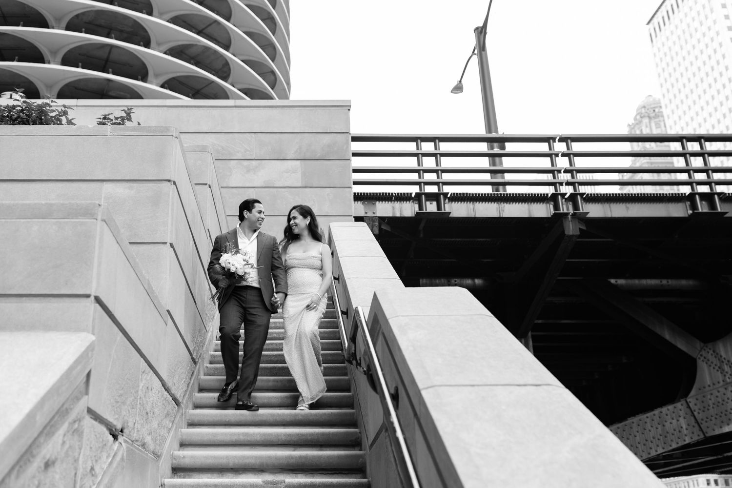 Black and white photo of bride and groom descending urban stairs with Chicago skyline in background.