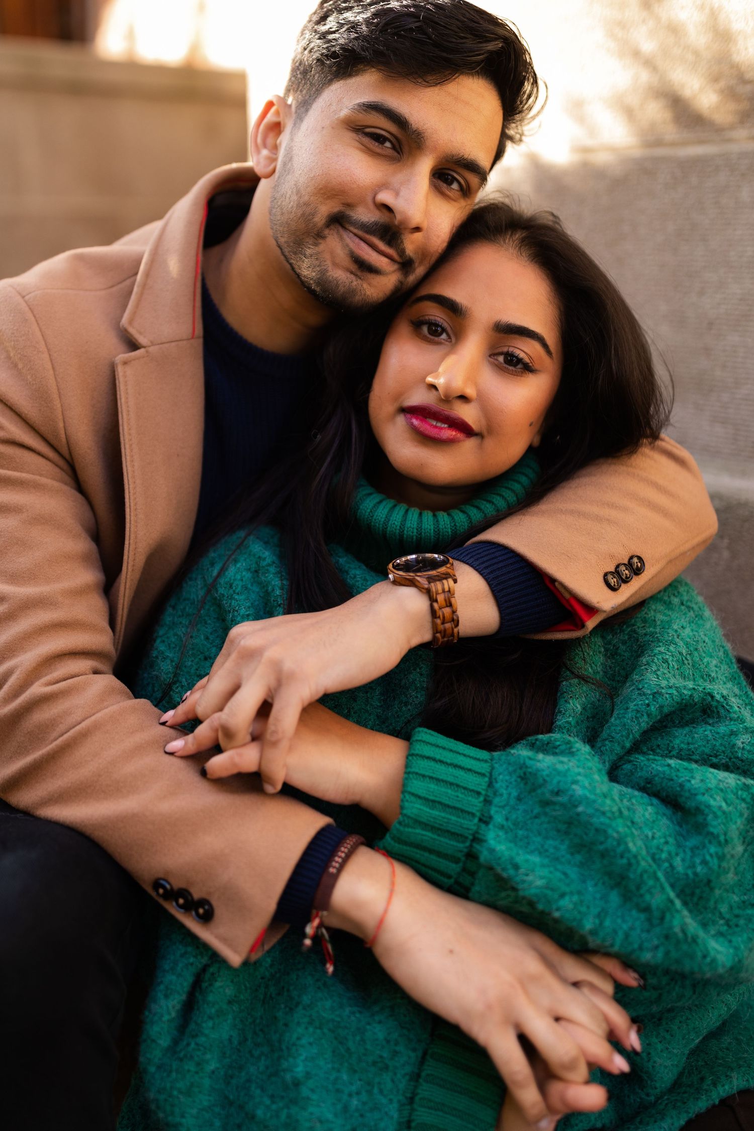 Couple embracing warmly, man in tan blazer hugging woman in teal sweater, wearing wooden watch, sitting outdoors in autumn.