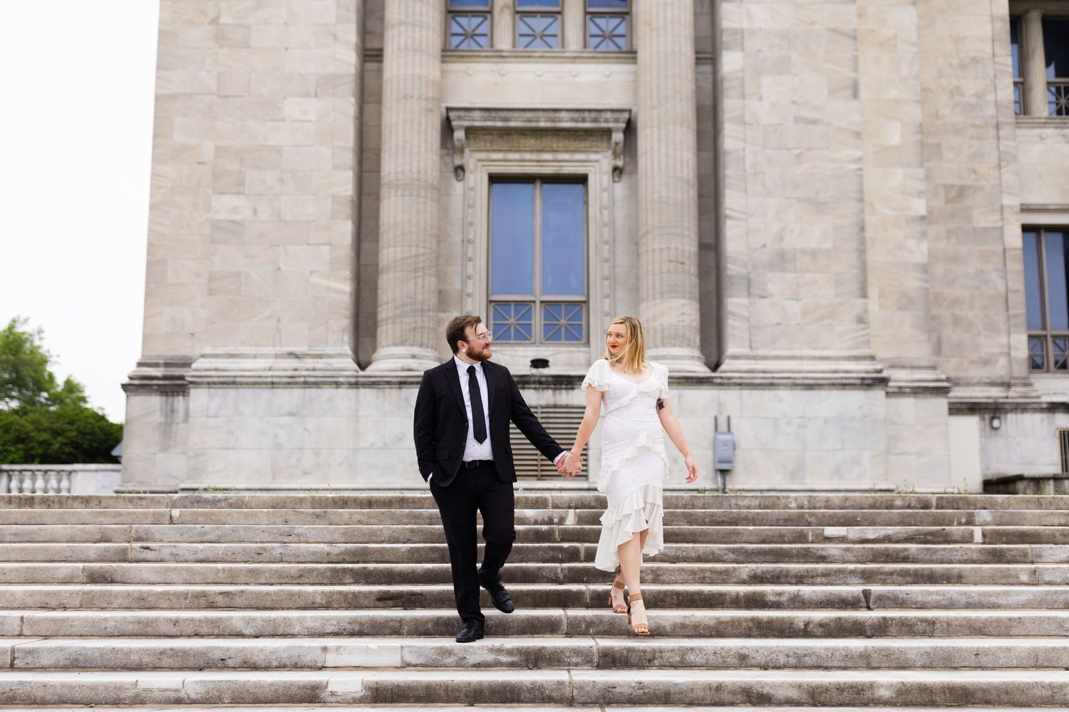 Bride and groom hold hands walking down stone steps of a grand neoclassical building.