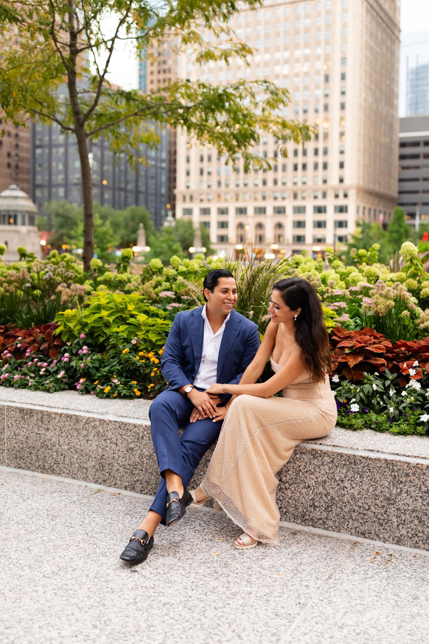 Engaged couple sitting on stone bench in urban garden, man in blue suit, woman in gold sequin gown.