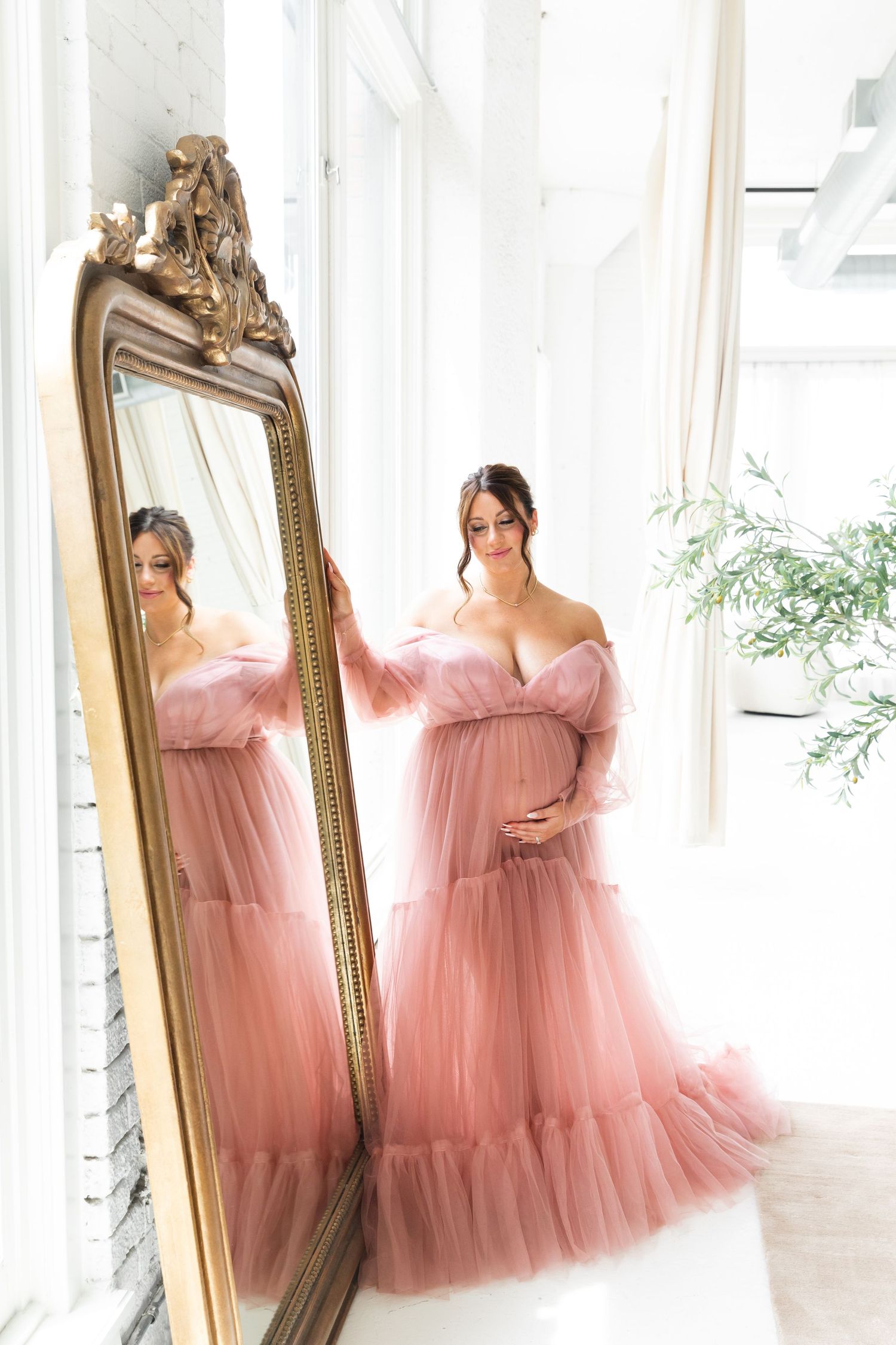 Pregnant woman in blush pink tulle maternity gown posing beside ornate gold mirror in bright studio.