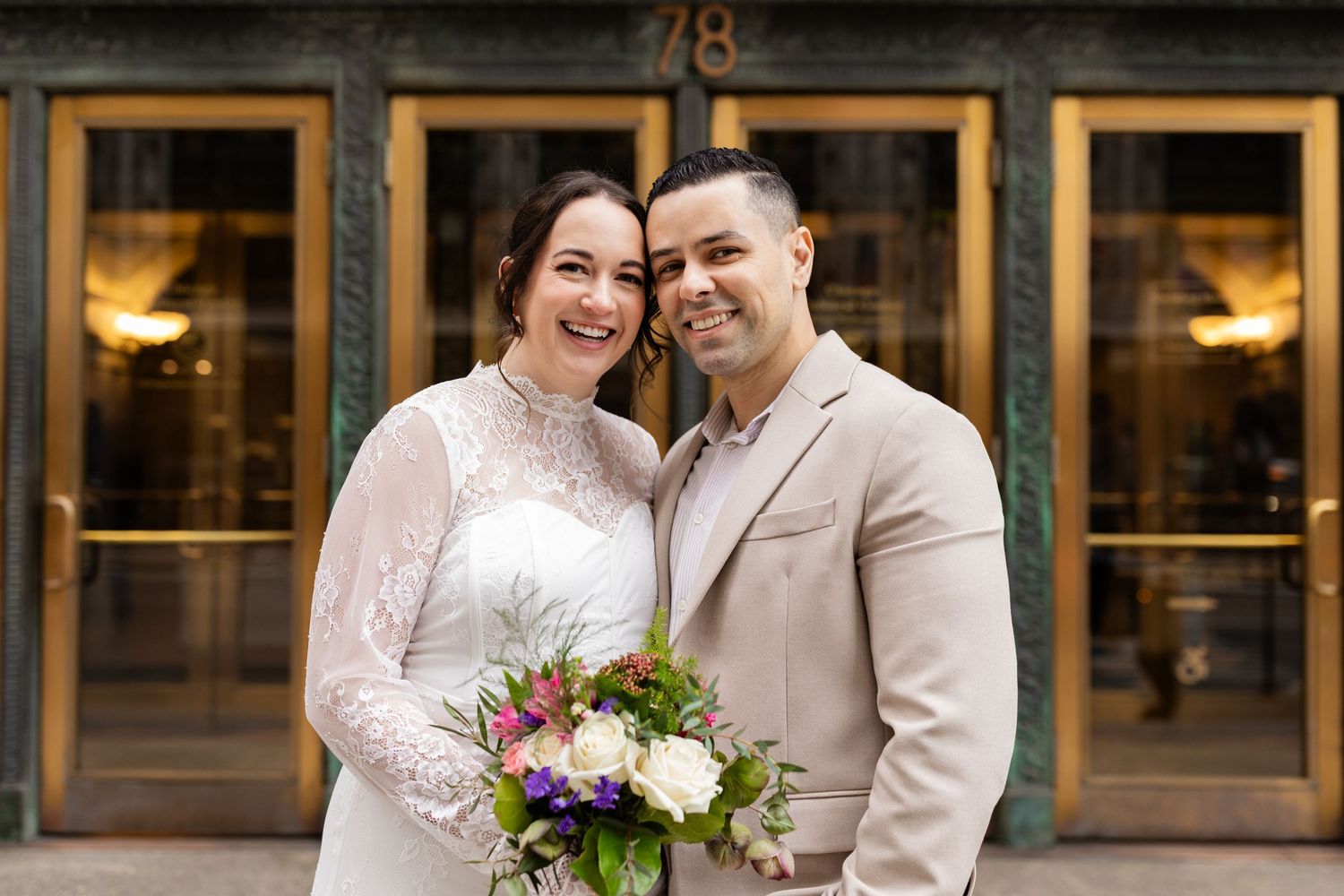 Smiling bride and groom posing together outside building 78, bride holding colorful wedding bouquet.