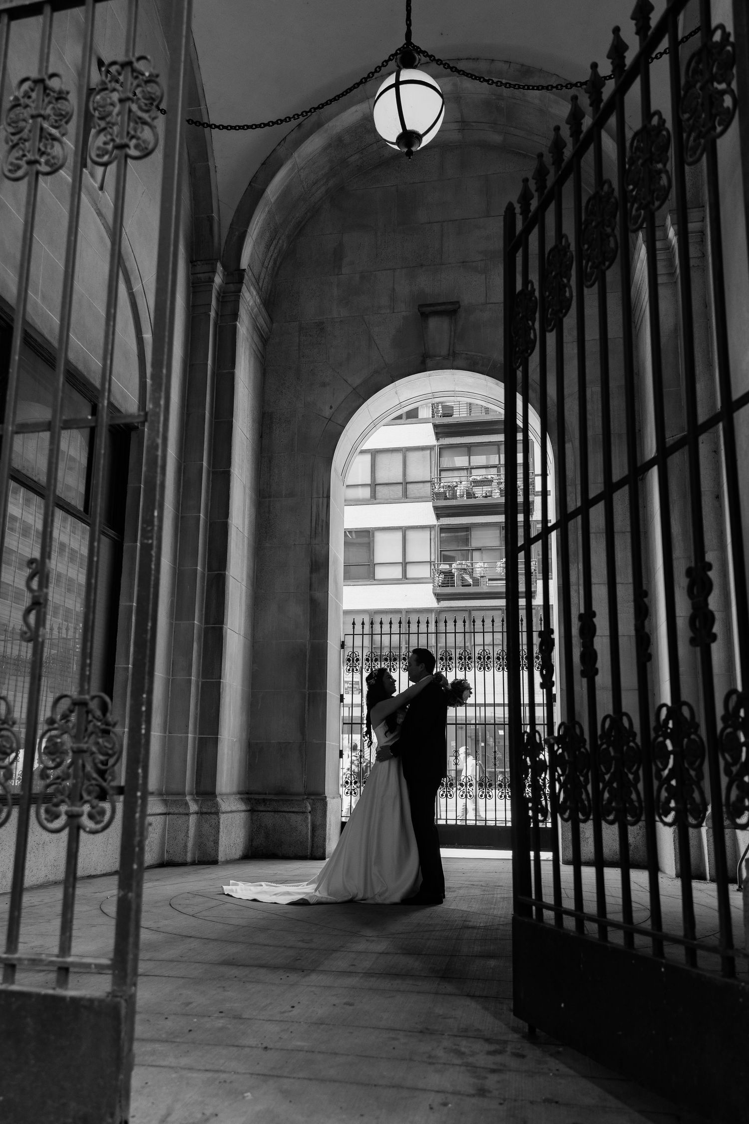 Black and white photo of a bride and groom sharing a romantic moment under an ornate arched iron gateway.