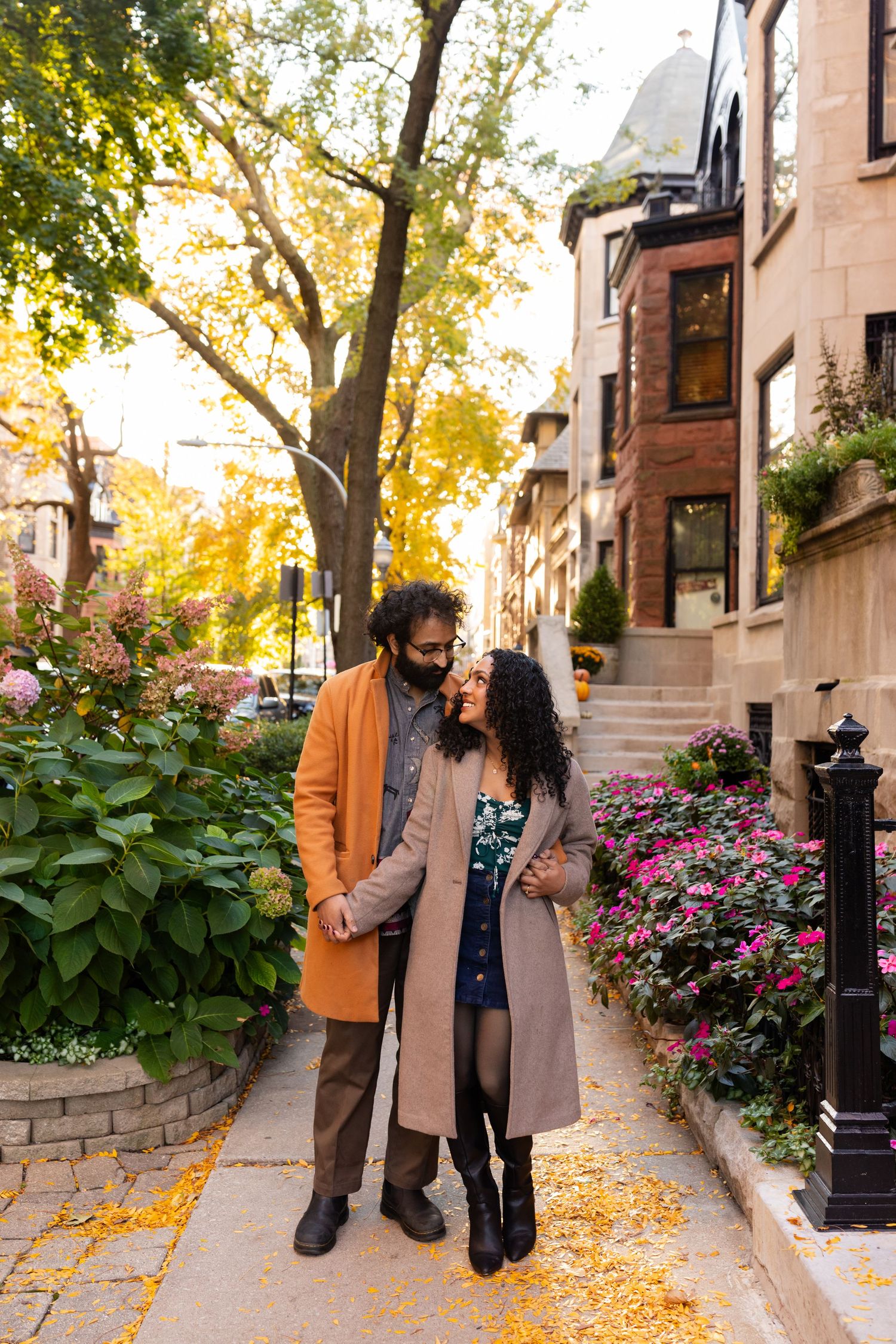 A couple shares a tender moment on a scenic autumn sidewalk lined with colorful flowers.