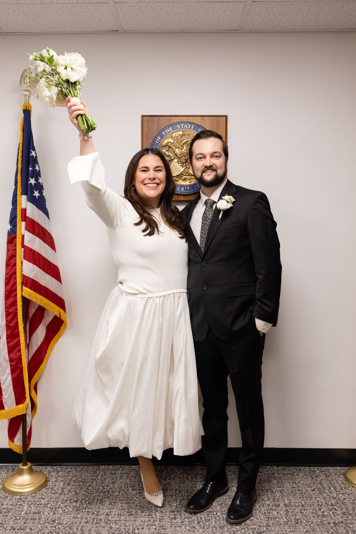 Jubilant bride raises bouquet in celebration beside groom in front of Illinois state seal and American flag.