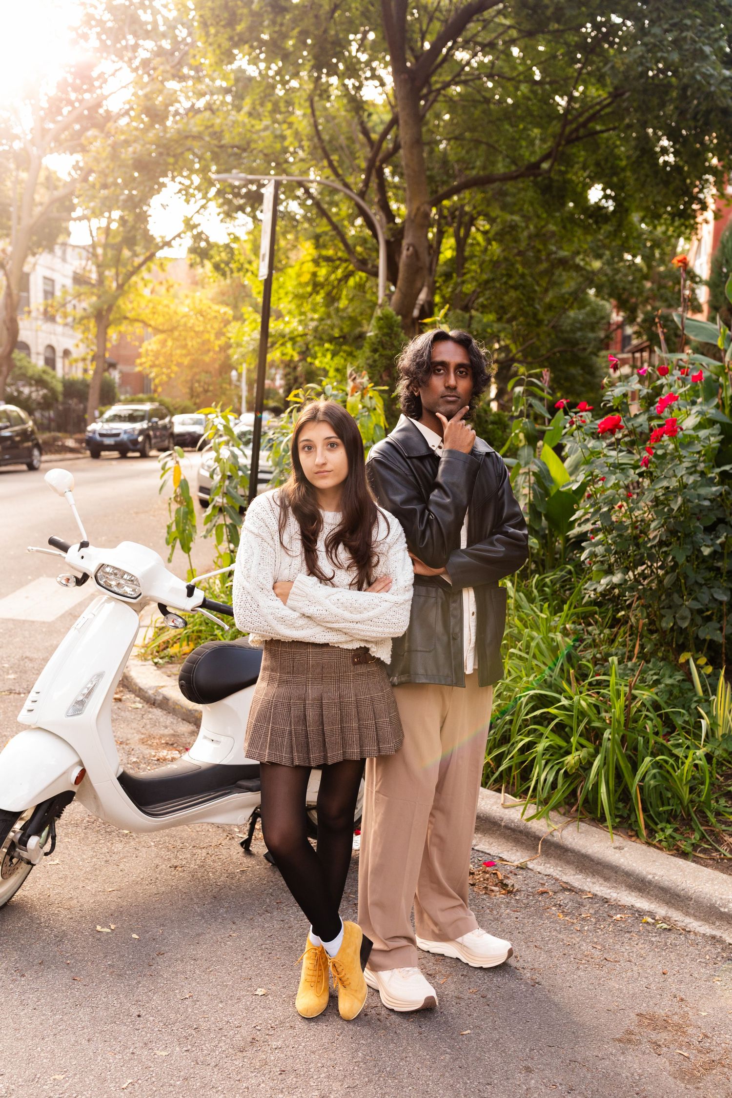 Stylish couple posing on urban street beside white scooter surrounded by greenery.