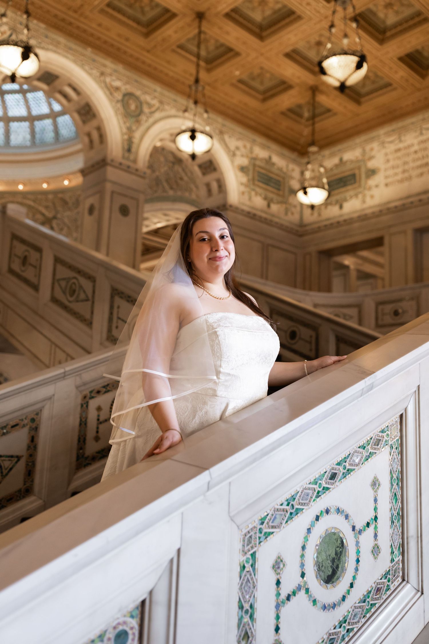 Bride in a lace wedding gown and veil posing on an ornate marble staircase in a grand historic building.