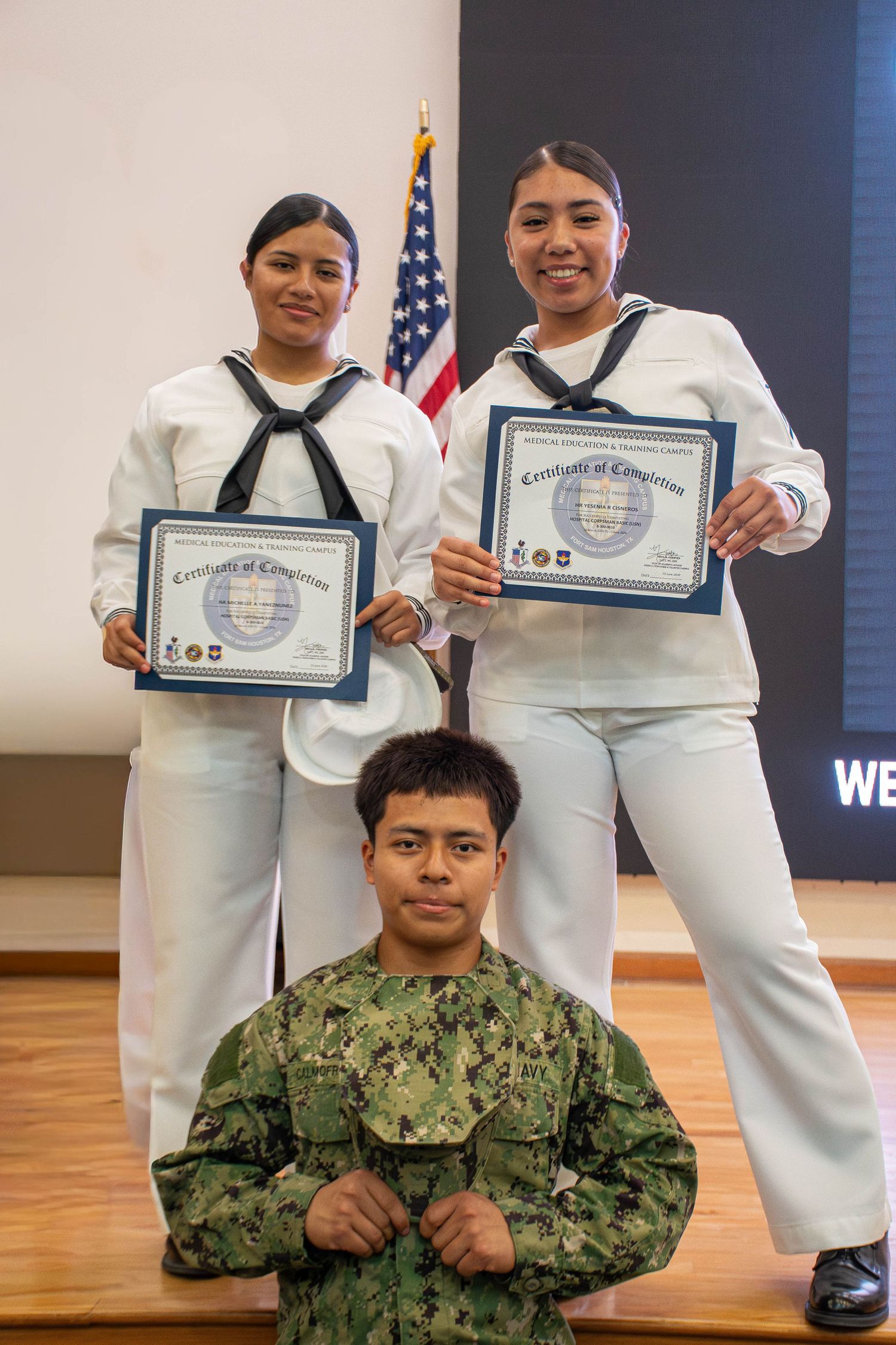 Three US Navy sailors posing with Certificates of Completion in front of American flag indoors.